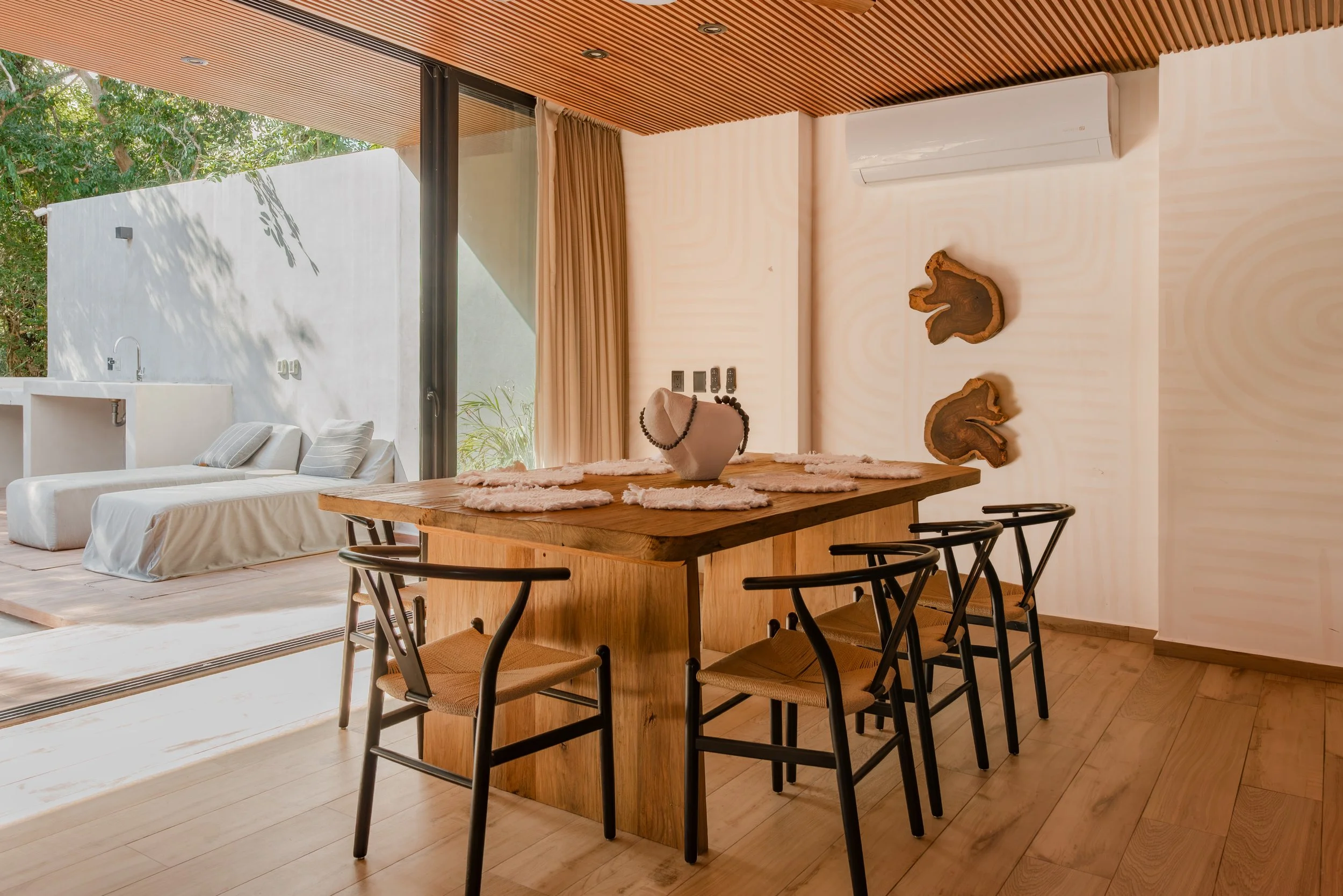 Modern interior of a room with a wooden ceiling, beige walls, and a wooden dining table with black chairs. A large sliding glass door opens to an outdoor area with trees. The room has a white sofa, decorative wooden wall art, a potted plant, and an air conditioner.