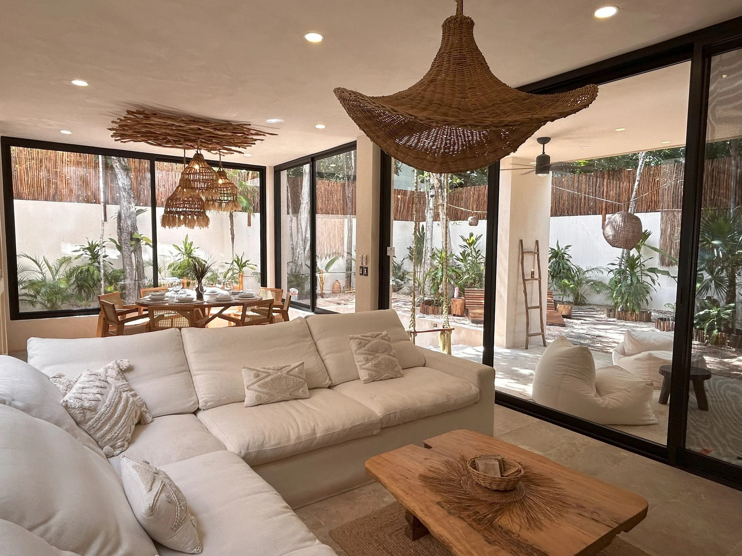 Living room with large white sectional sofa, wooden coffee table, decorative pillows, surrounded by large glass windows showing outdoor greenery and garden area, with woven pendant lights hanging from ceiling.