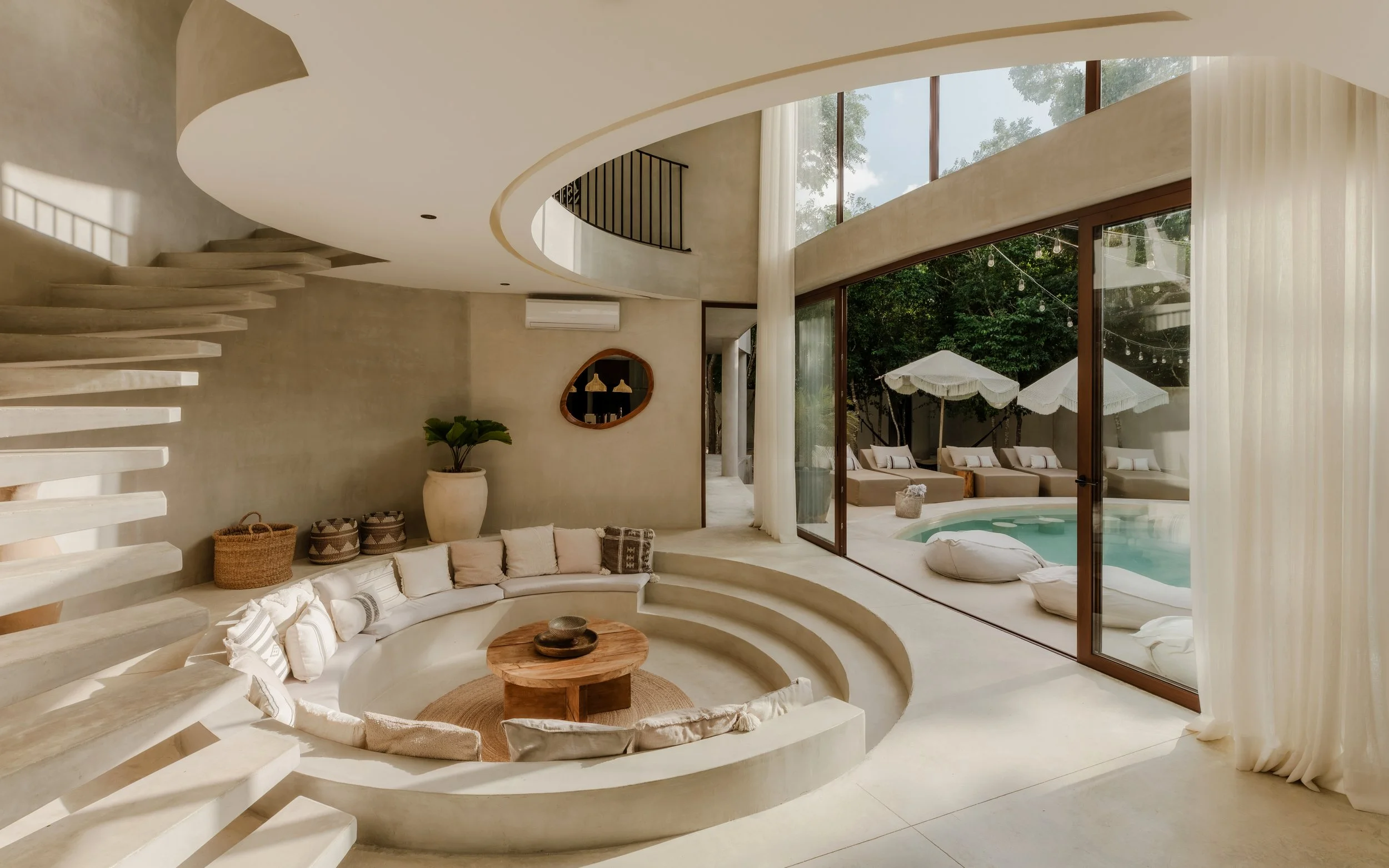 Modern living room with a built-in curved sofa, round wooden coffee table, and large glass windows overlooking a patio with umbrellas and greenery.