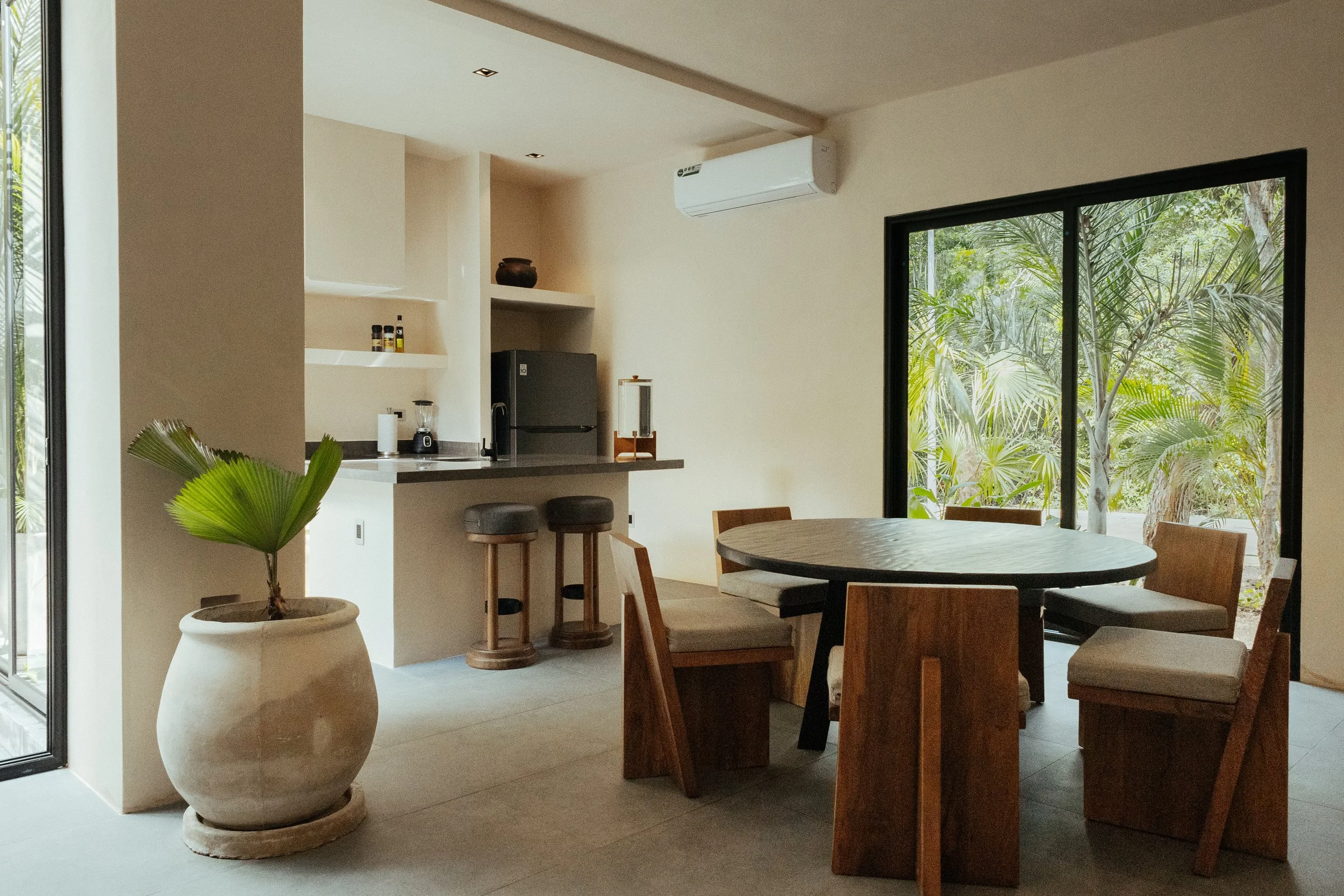 Interior view of a dining area with a round wooden table, wooden chairs, large windows showing greenery, and a kitchen area with a gray refrigerator, open shelves, and countertop with appliances and decor.