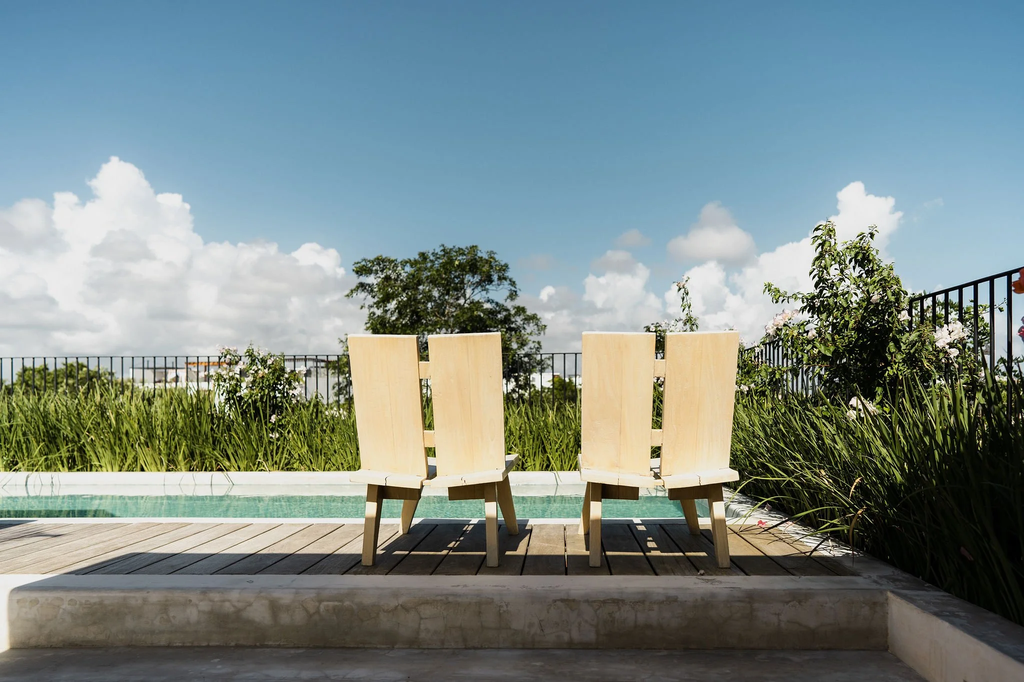 Three wooden deck chairs on a patio facing outside with greenery and a tree in the background under a blue sky with scattered clouds.