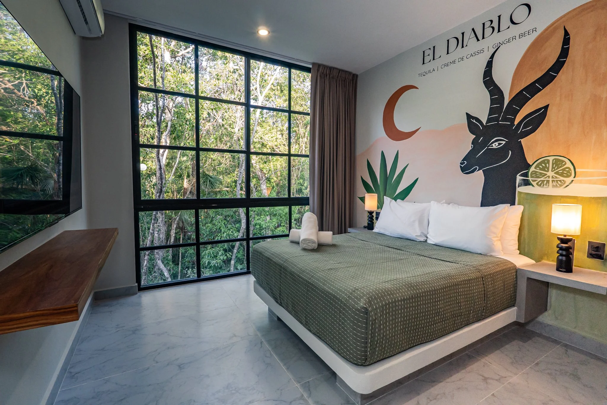 A bedroom with a large window showing greenery outside, a mural of a blacked antelope with horns, a green plant, a bed with white and green bedding, and a nightstand with a lamp.