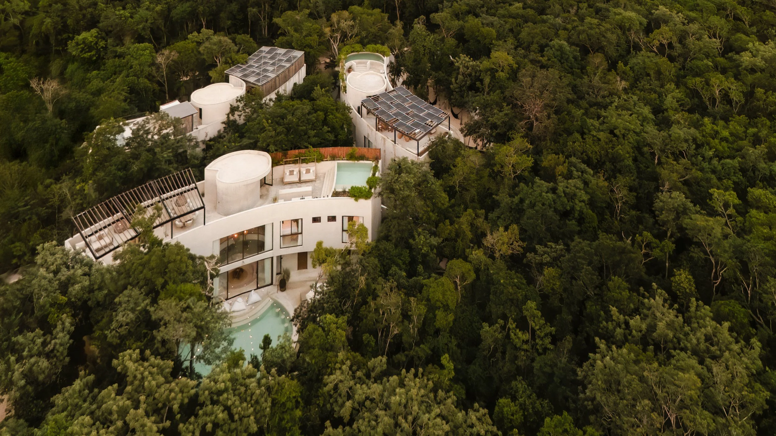 Aerial view of a modern white building with rooftop pools, solar panels, and outdoor seating, surrounded by dense green forest.