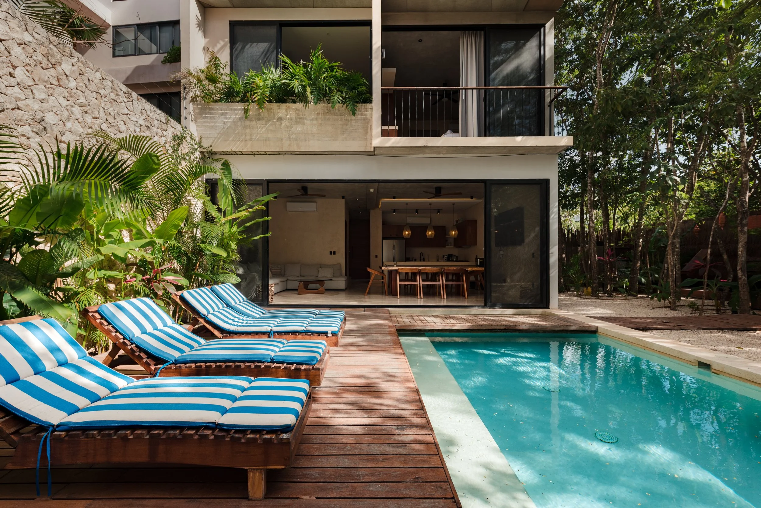 A modern house with large glass sliding doors opening to a swimming pool and outdoor lounge area with striped blue and white chairs. There's lush green foliage and trees surrounding the backyard, creating a private and tropical atmosphere.