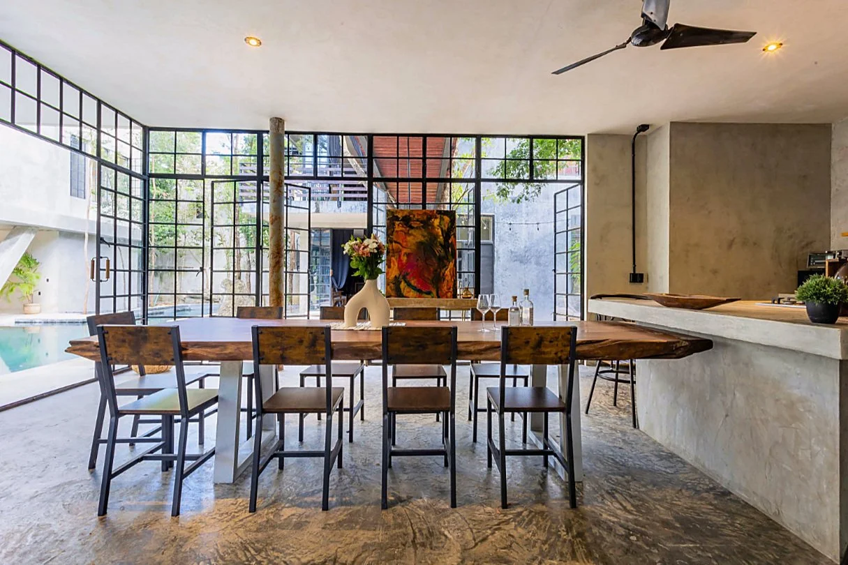 Modern dining area with a large wooden table, eight chairs, a vase of flowers, wine glasses, a painting, and large glass windows with a view of an outdoor patio and pool.