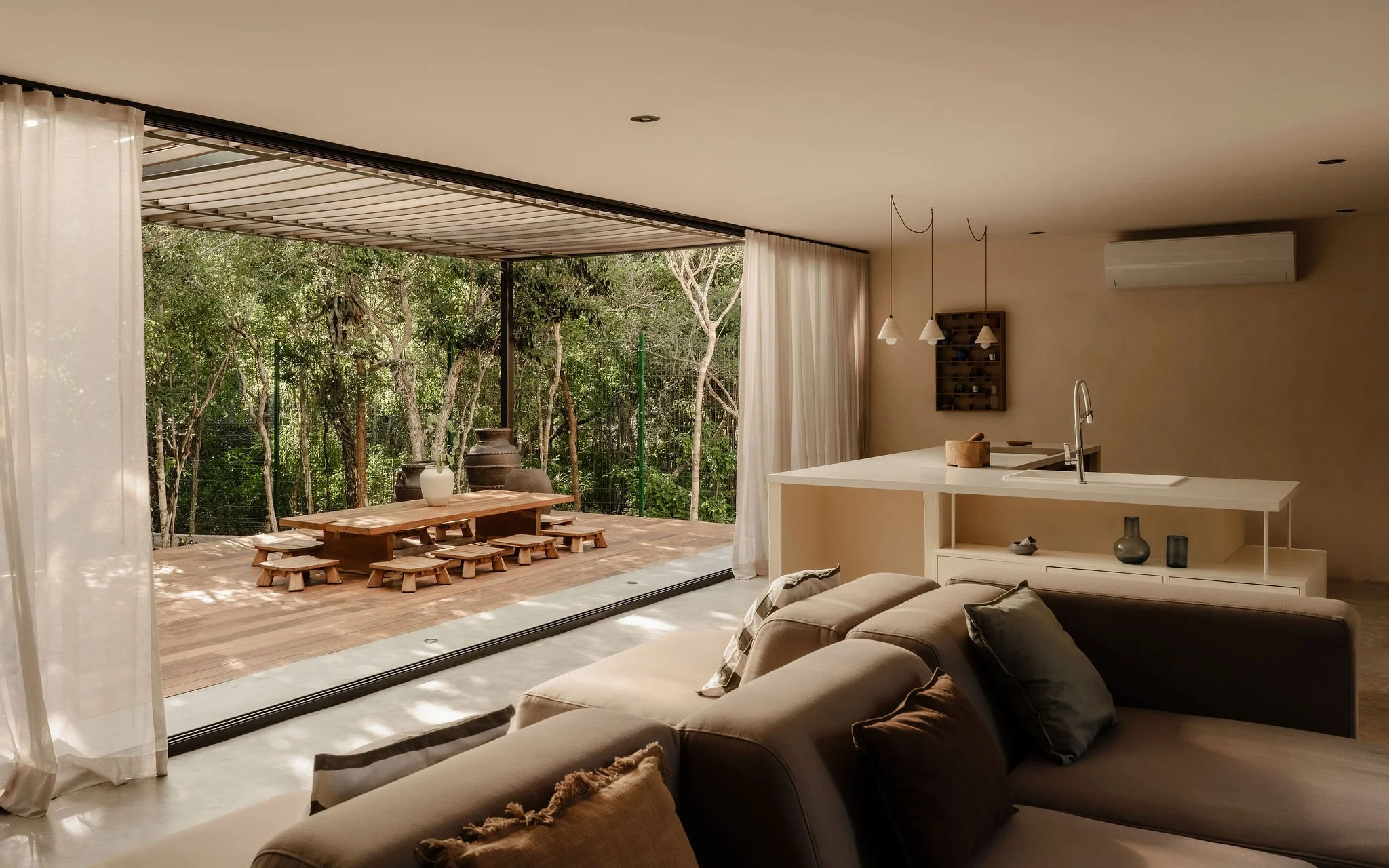 Modern living room with large sliding glass doors opening to a lush outdoor patio with wooden table and benches, overlooking green trees.