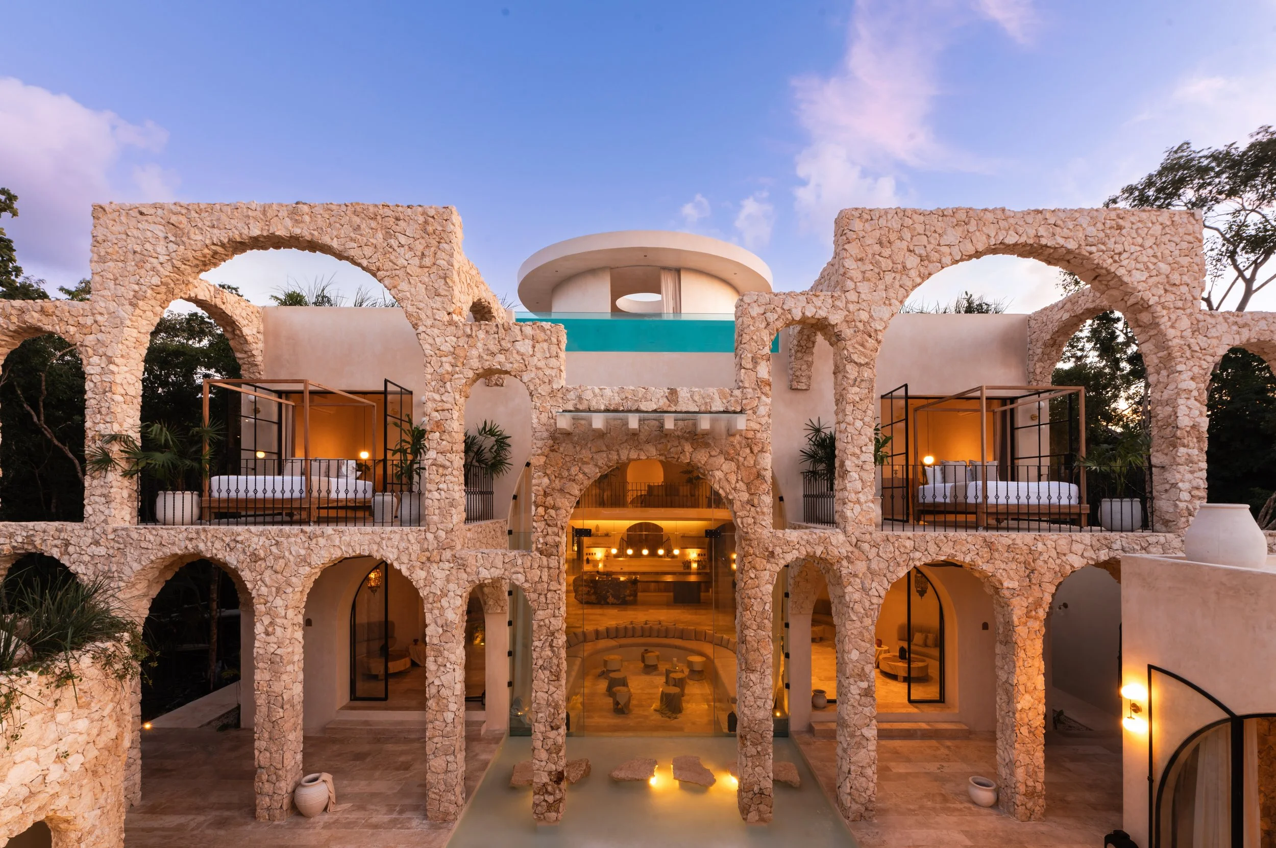 A multi-story modern building with stone arches, open balconies, and a rooftop pool, illuminated at dusk with a clear sky overhead.