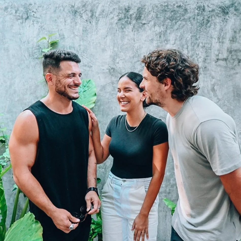 Three friends standing outdoors against a concrete wall with green plants, smiling and enjoying a conversation.