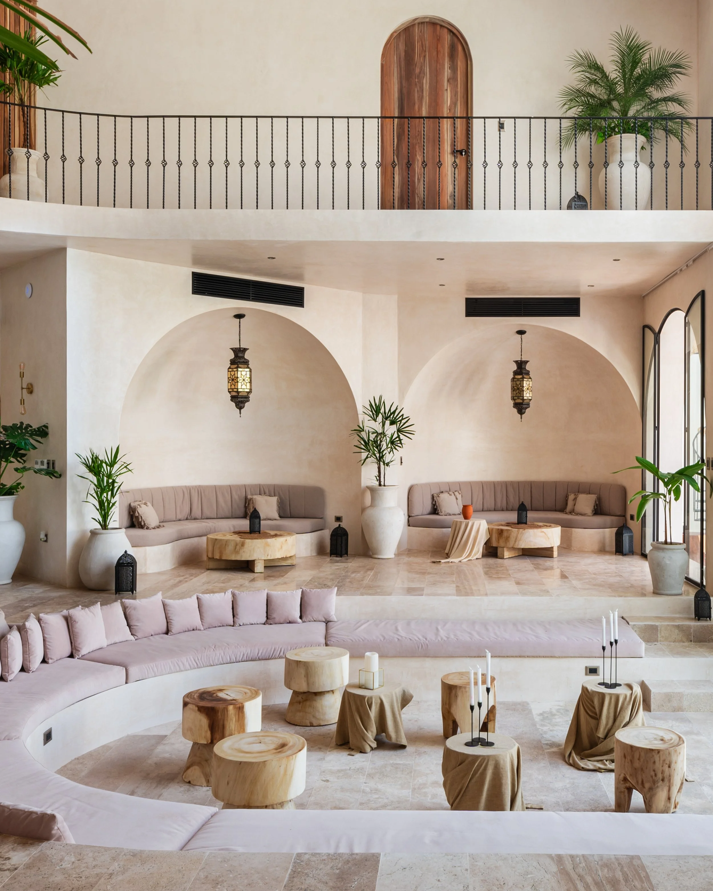 Elegant lobby with curved beige seating, wooden stools, and small tables draped in beige cloths. Green potted plants in white vases and black lanterns add decor. Archways and hanging lanterns highlight a Middle Eastern or Mediterranean style.