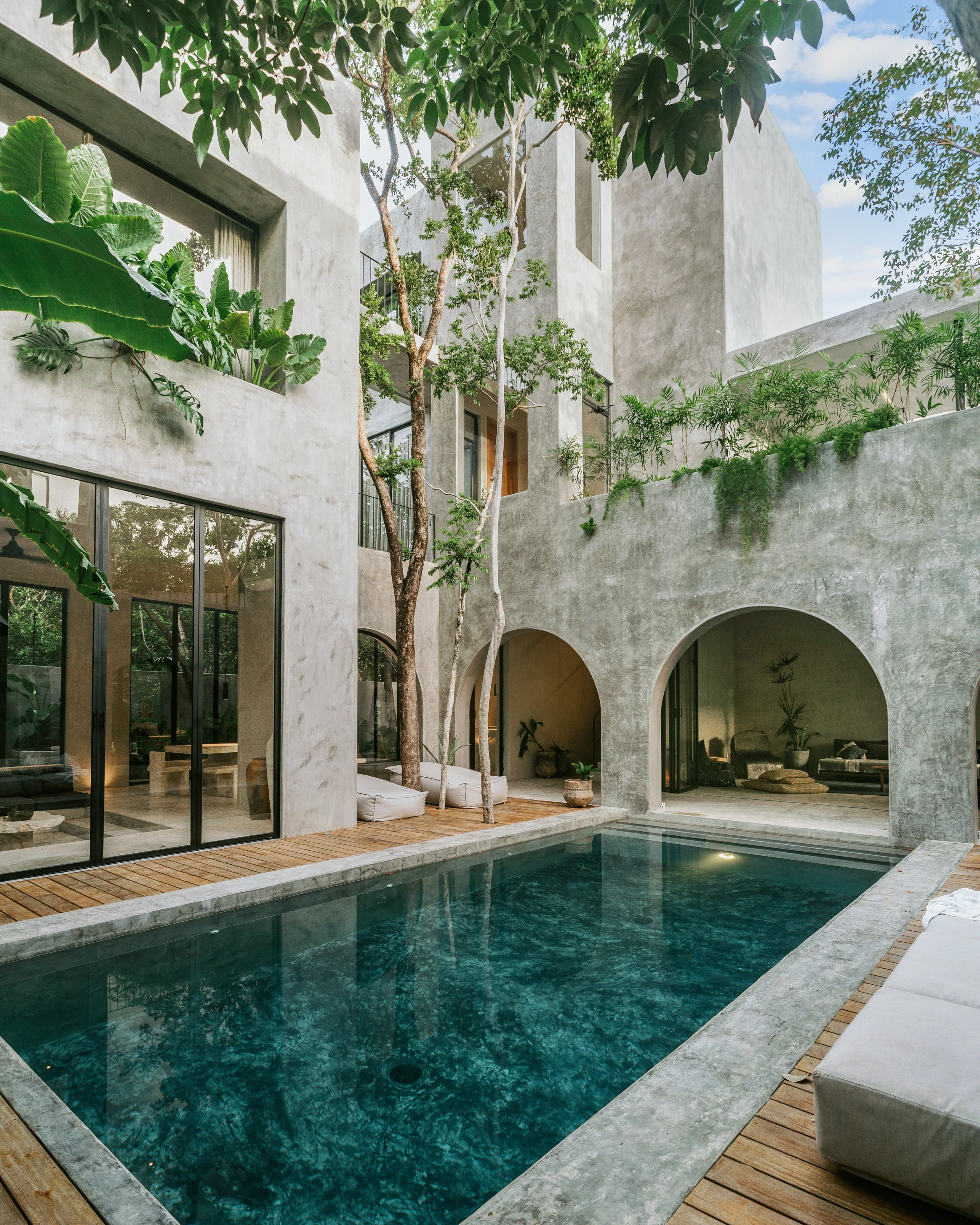 Modern courtyard with a swimming pool surrounded by concrete walls, green plants, and trees, with outdoor seating and large windows.