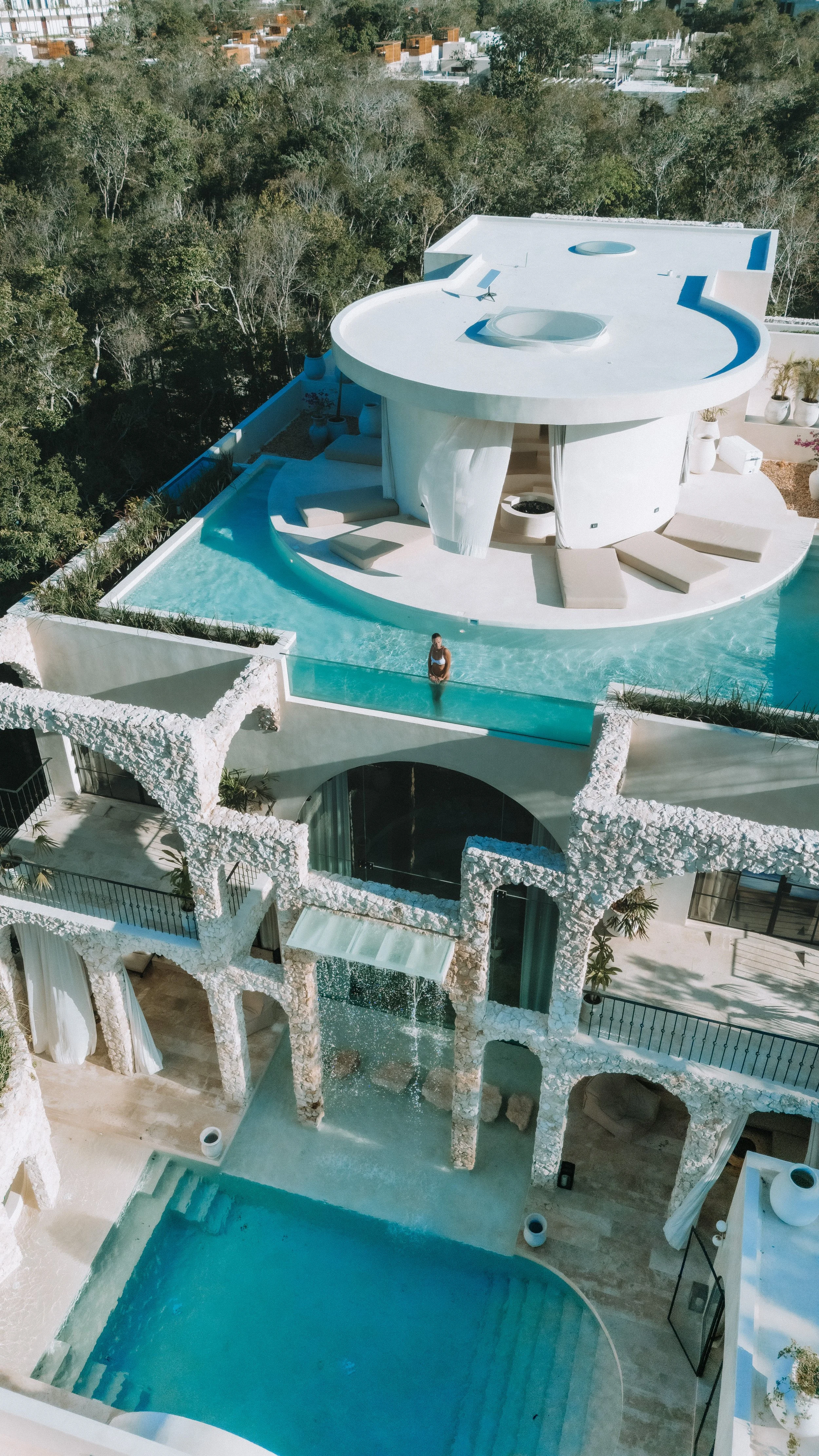 Aerial view of a modern white house with an infinity pool, outdoor seating area with beige cushions, surrounded by trees, with additional structures in the background.