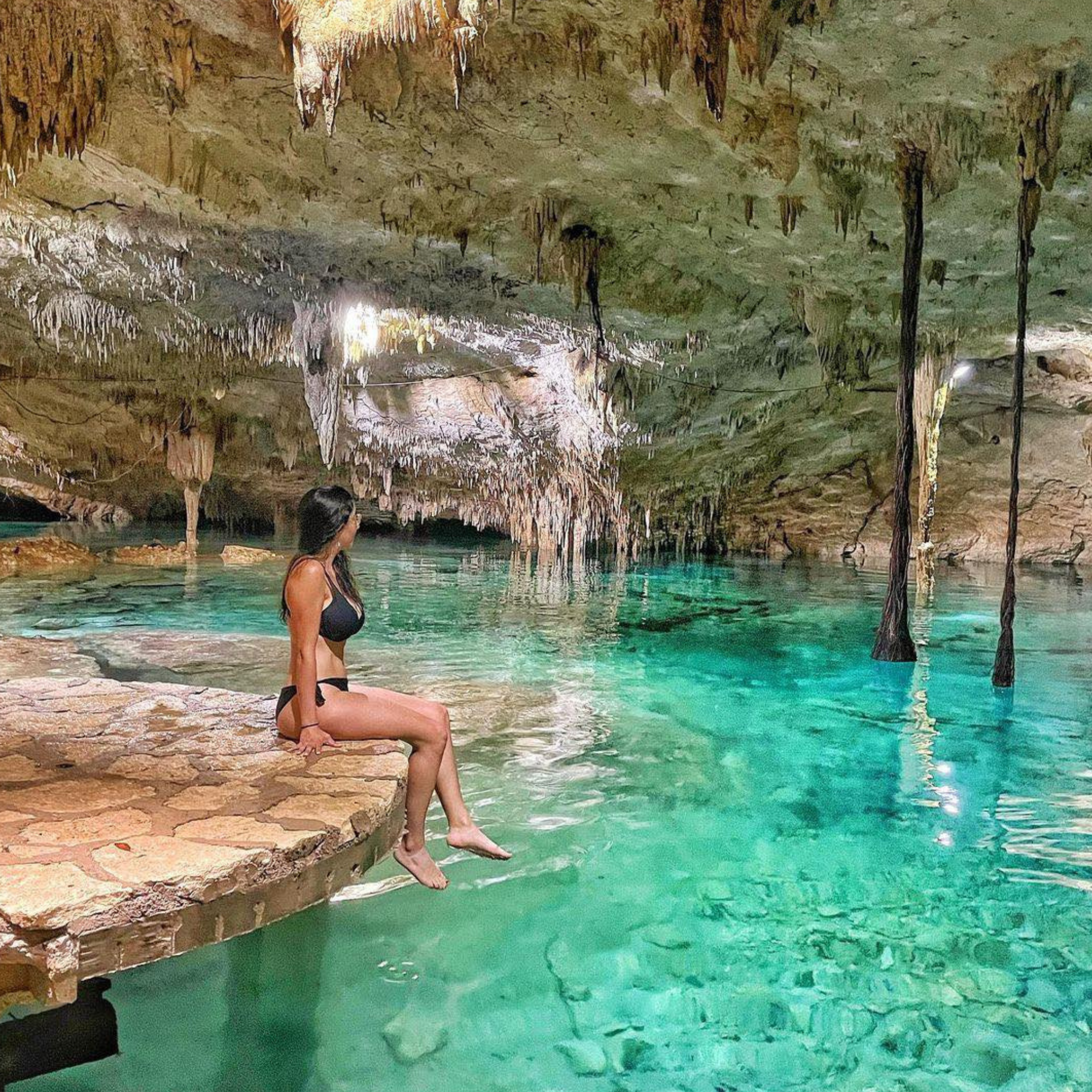 A woman in a black bikini sitting on rocks inside a cave with turquoise water and stalactites hanging from the ceiling.