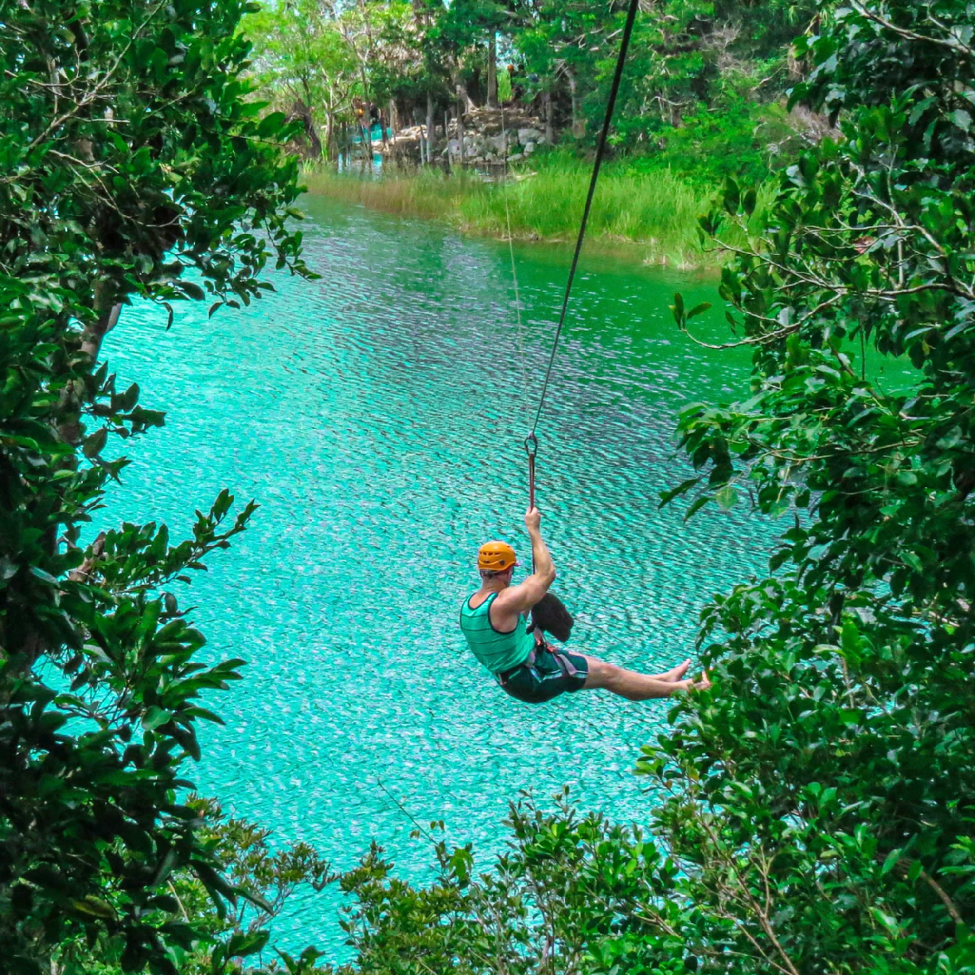 A person wearing a helmet and blue tank top is sitting on the edge of the water, holding onto a swing hanging from a tree branch over a turquoise river, surrounded by lush green foliage.