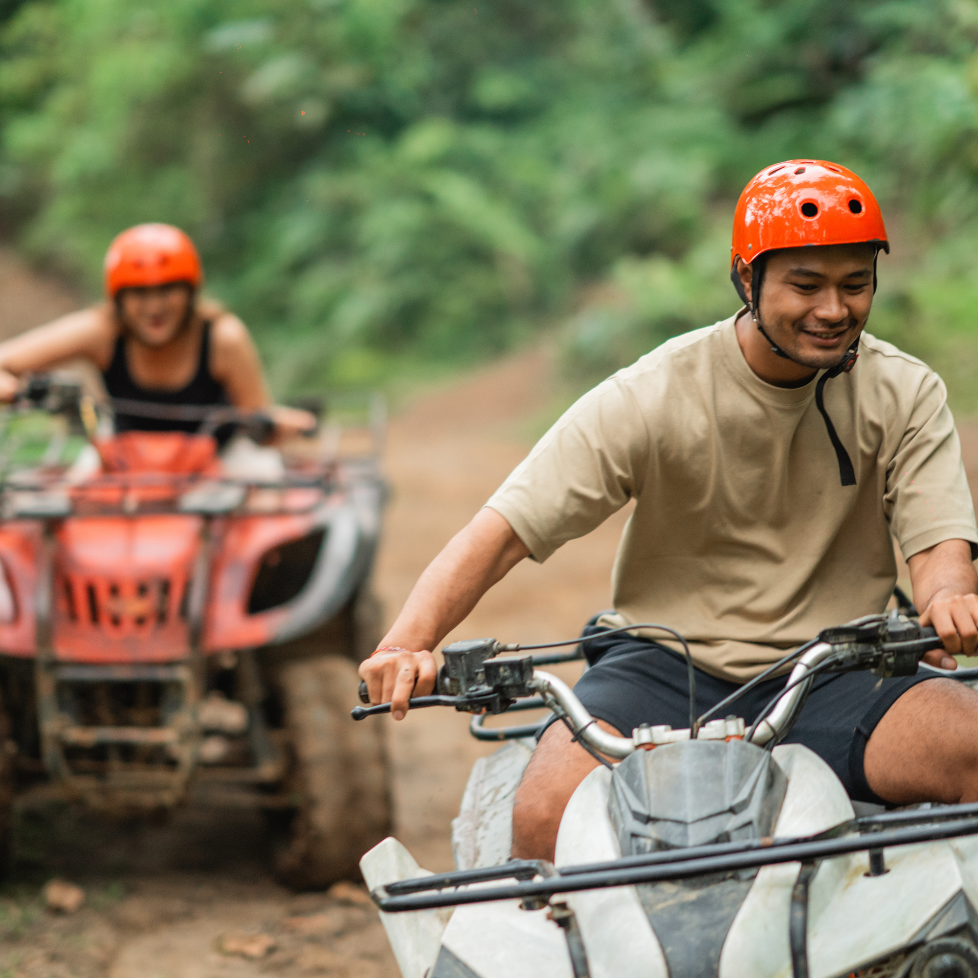 Two people riding motorcycles on a dirt trail in a wooded area, both wearing orange helmets and casual clothing, with the focus on the person in the foreground.