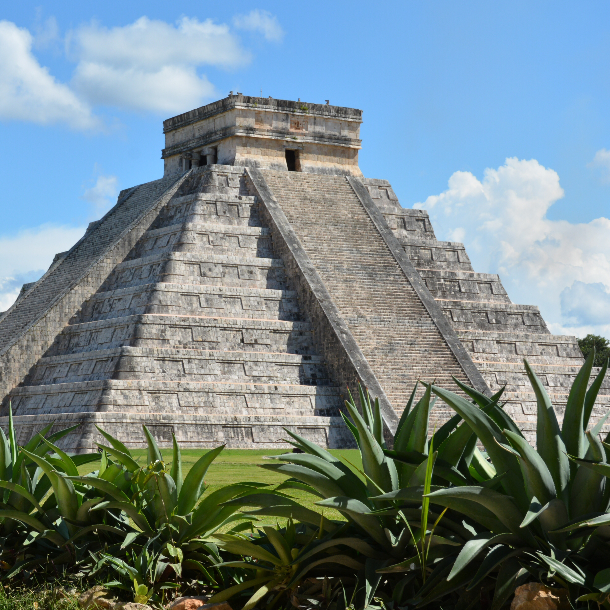 Ancient stone pyramid with tiered steps, surrounded by green plants and a bright sky with white clouds.