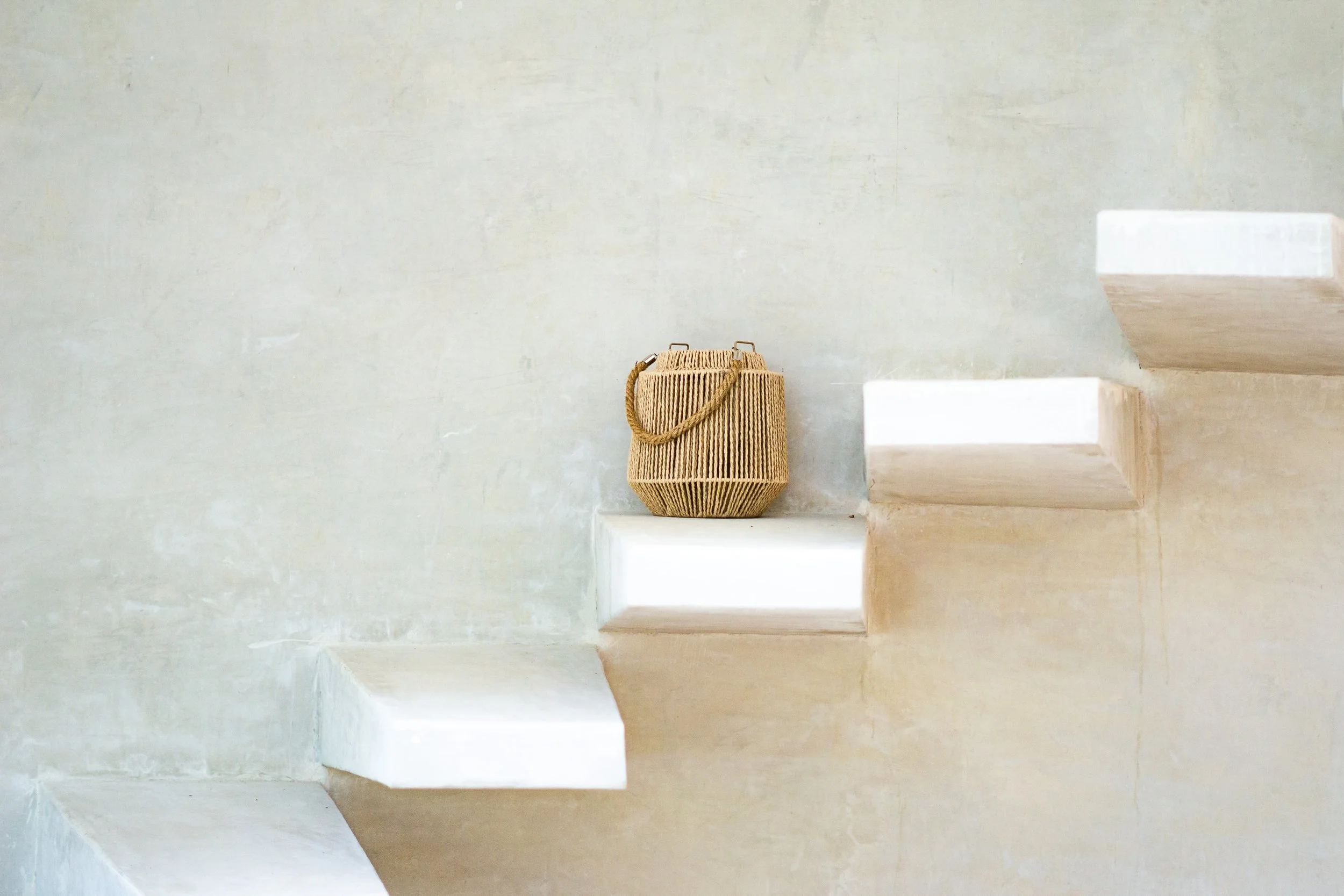 A wicker handbag with a handles, placed on a white marble staircase against a plain light-colored wall.