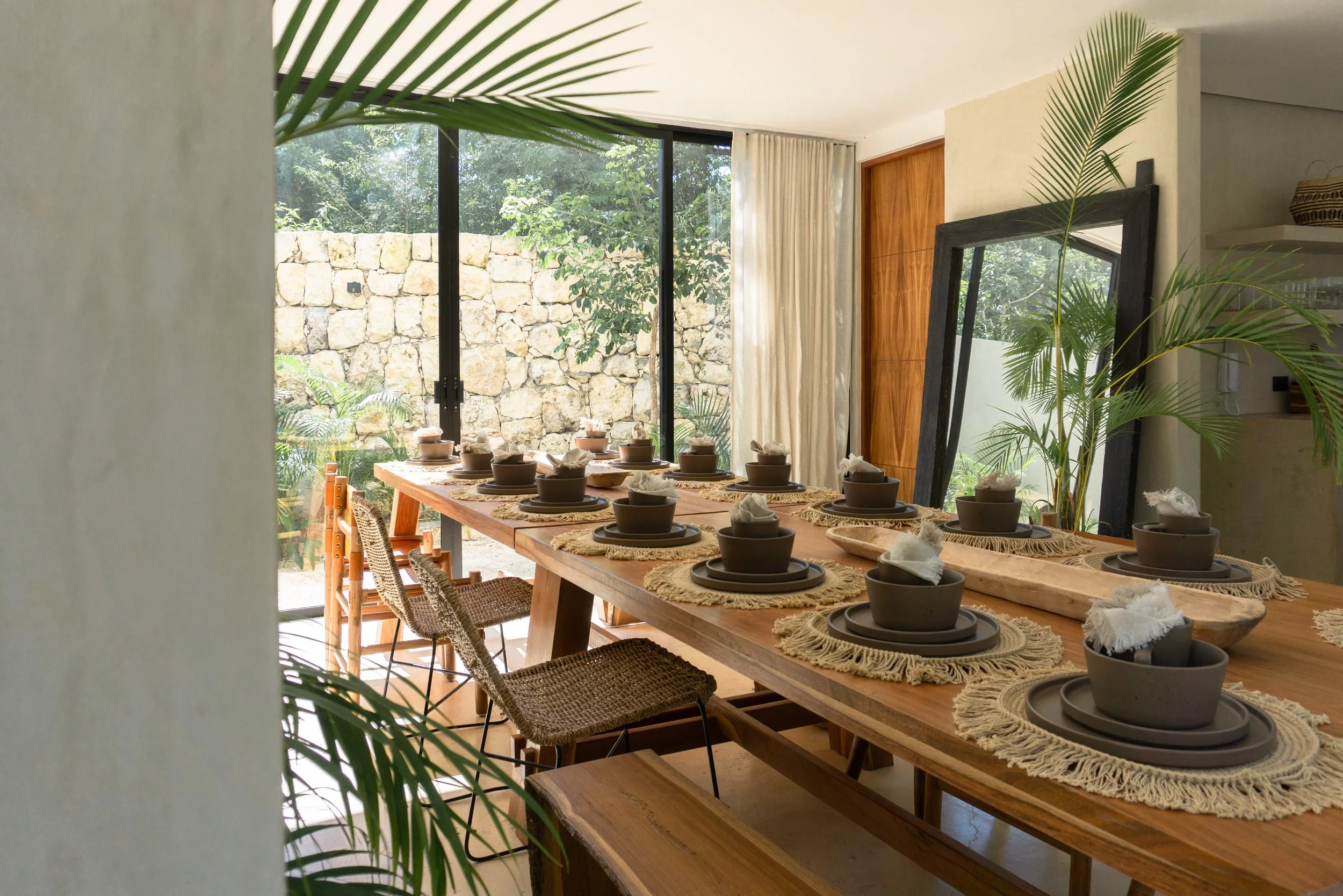 A dining table set with bowls, plates, and cloth napkins inside a bright room with a large window revealing outdoor greenery and stone wall, decorated with plants and a large mirror.
