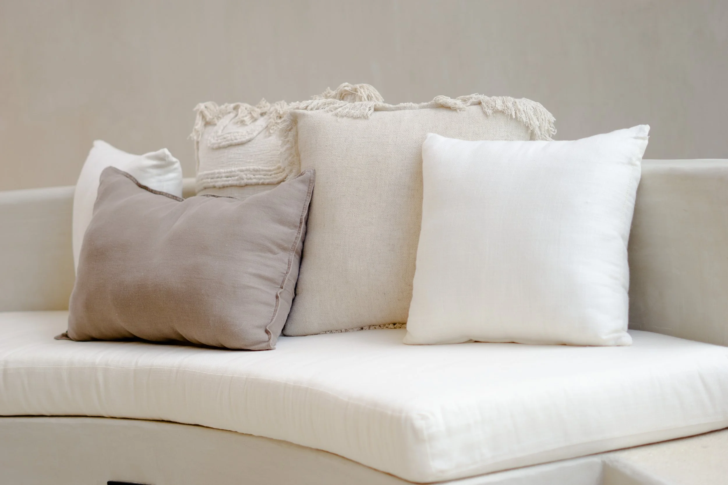 A collection of neutral-colored throw pillows on a white sofa, with a textured beige pillow in the center and plain white and light gray pillows on each side, against a beige wall.