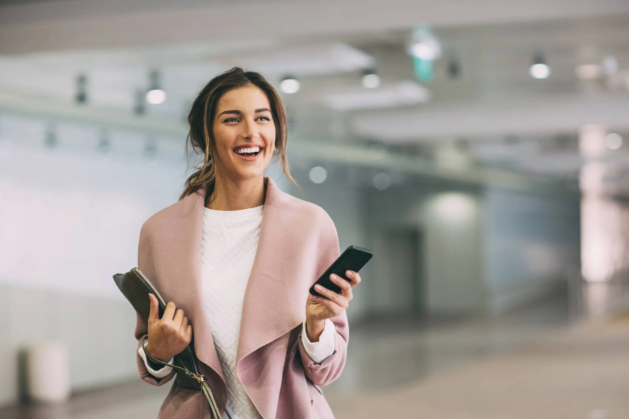 A woman holding a smartphone and a notepad, smiling in an indoor setting with blurred background.