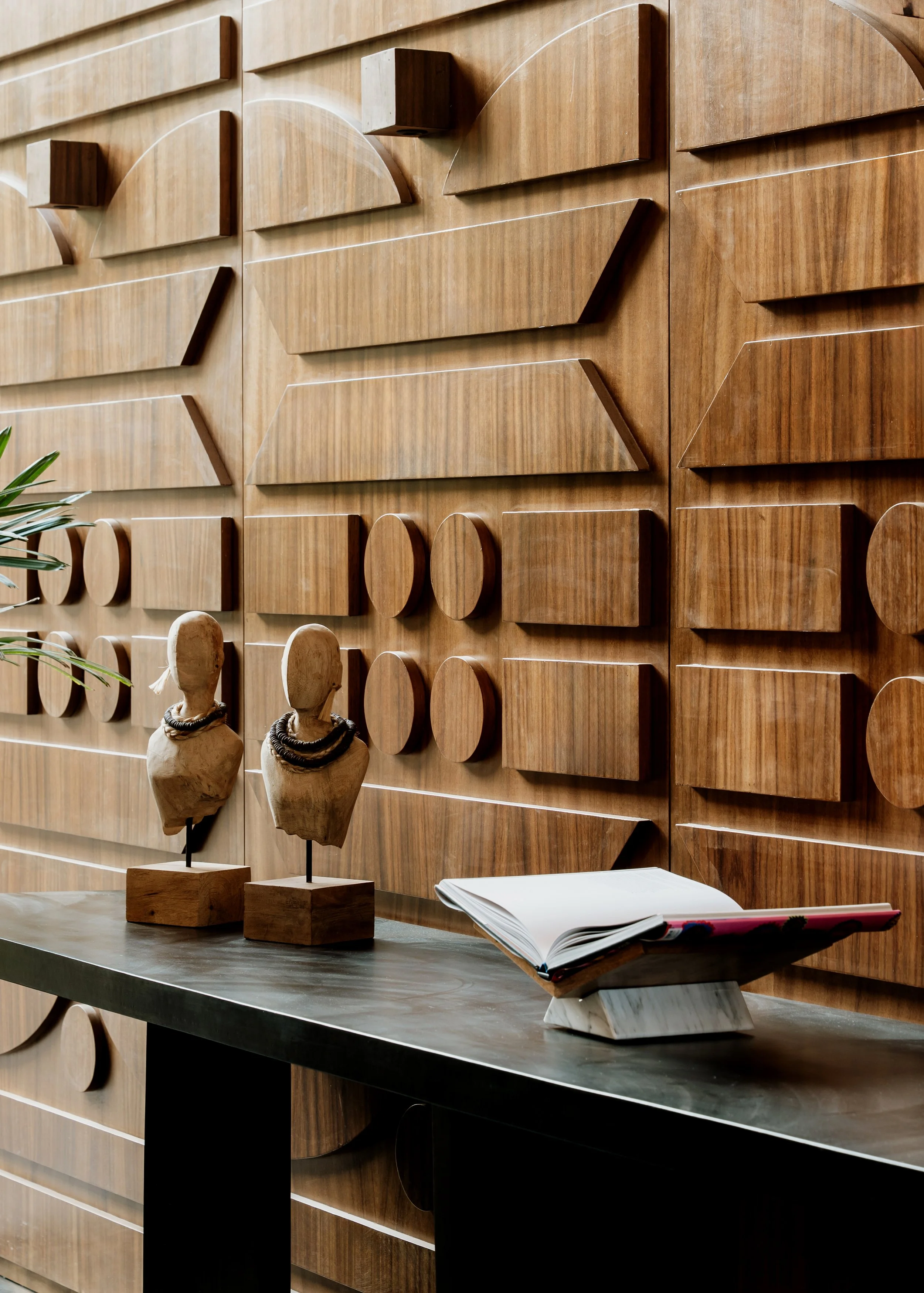 Decorative wooden wall with geometric patterns, two artist bust sculptures with necklaces on a table, and an open book with a pink pen on a marble stand.
