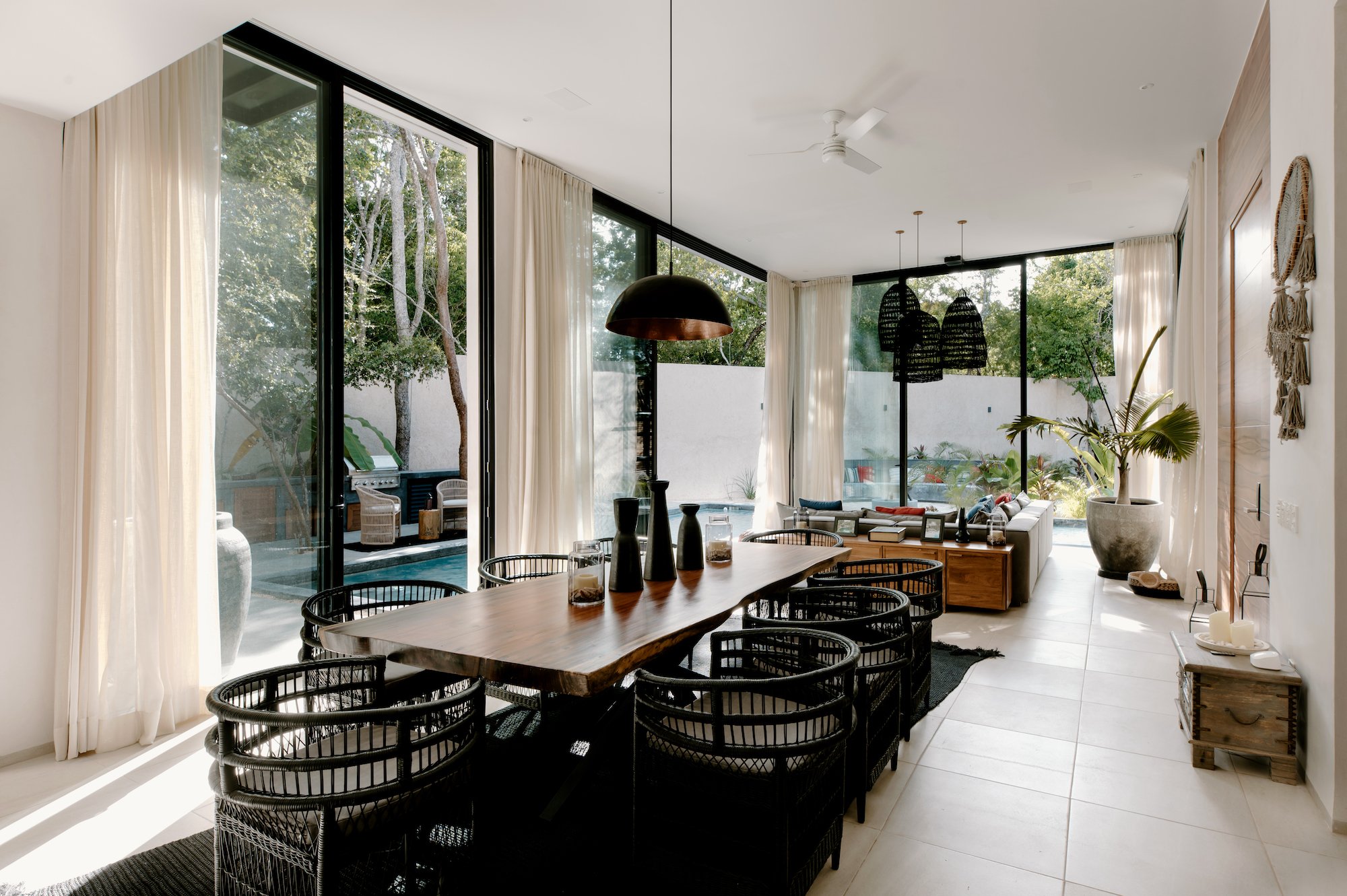 Bright, modern living room with large glass doors opening to outdoor area, white curtains, black wicker dining chairs around a wooden table, potted plants, hanging light fixtures, and a ceiling fan.