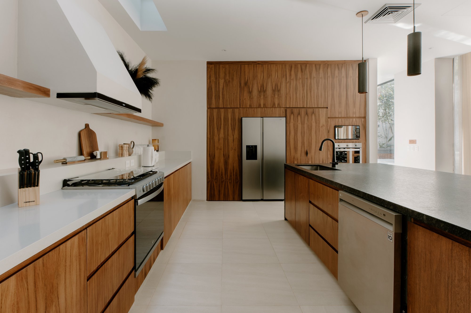 Modern kitchen with wooden cabinets, white countertops, and stainless steel appliances. There are minimalistic hanging lights and a window with natural light. A refrigerator and microwave are built into the wooden cabinetry.