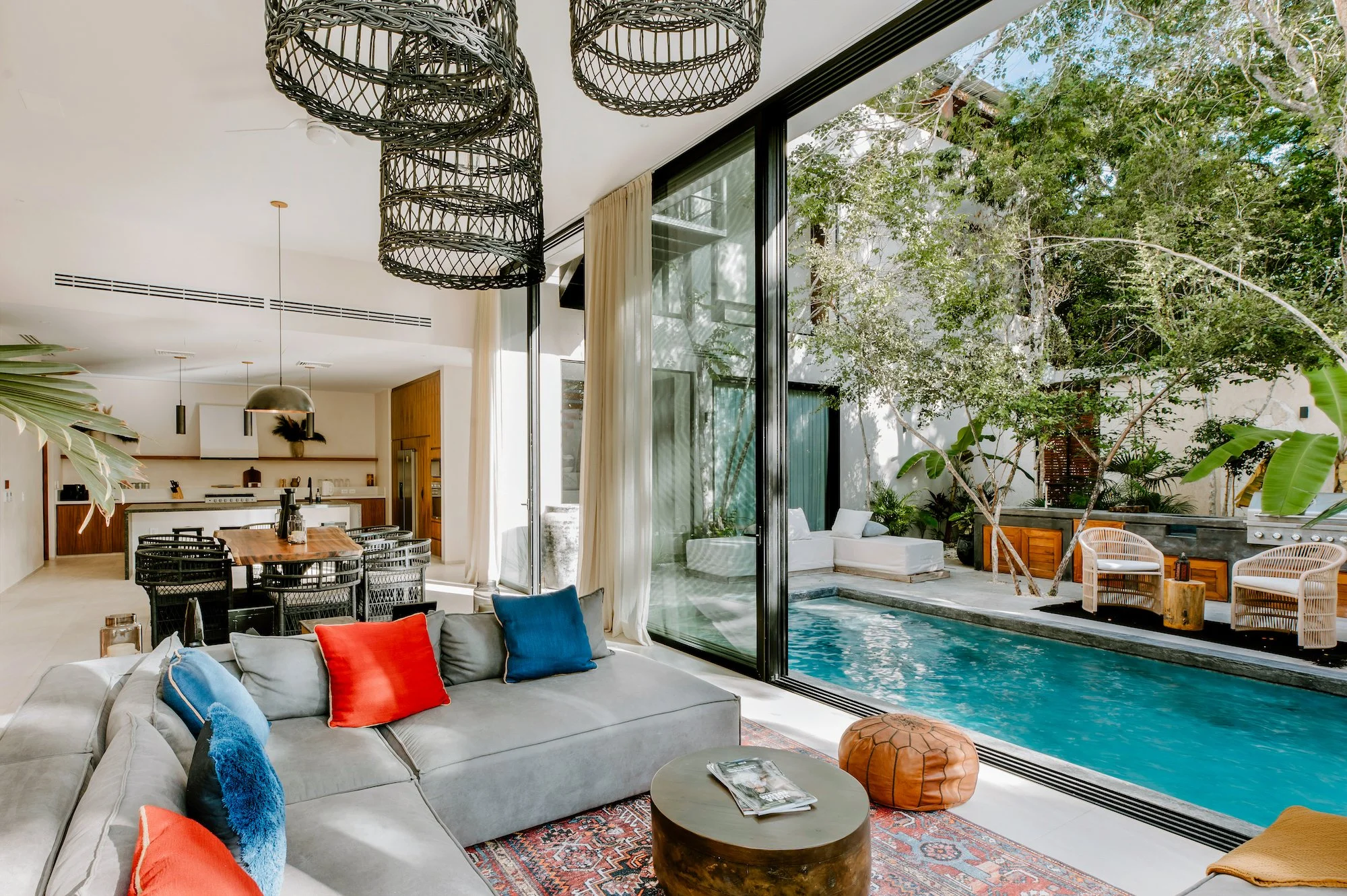 A modern indoor living area with a large L-shaped gray sofa decorated with red and blue pillows, a round wooden coffee table, a colorful patterned rug, and floor-to-ceiling glass doors opening to an outdoor pool area surrounded by lush green trees.