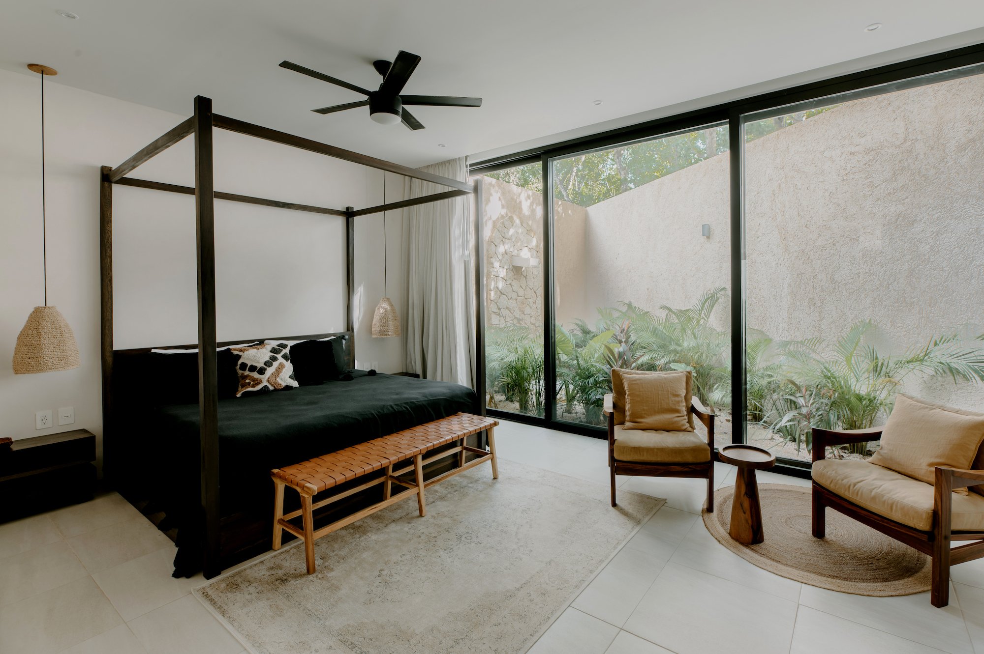 Modern bedroom with large glass sliding doors leading to outdoor greenery, featuring a black canopy bed with decorative pillows, wooden bench, small side tables, hanging pendant lamp, ceiling fan, and cozy armchairs.