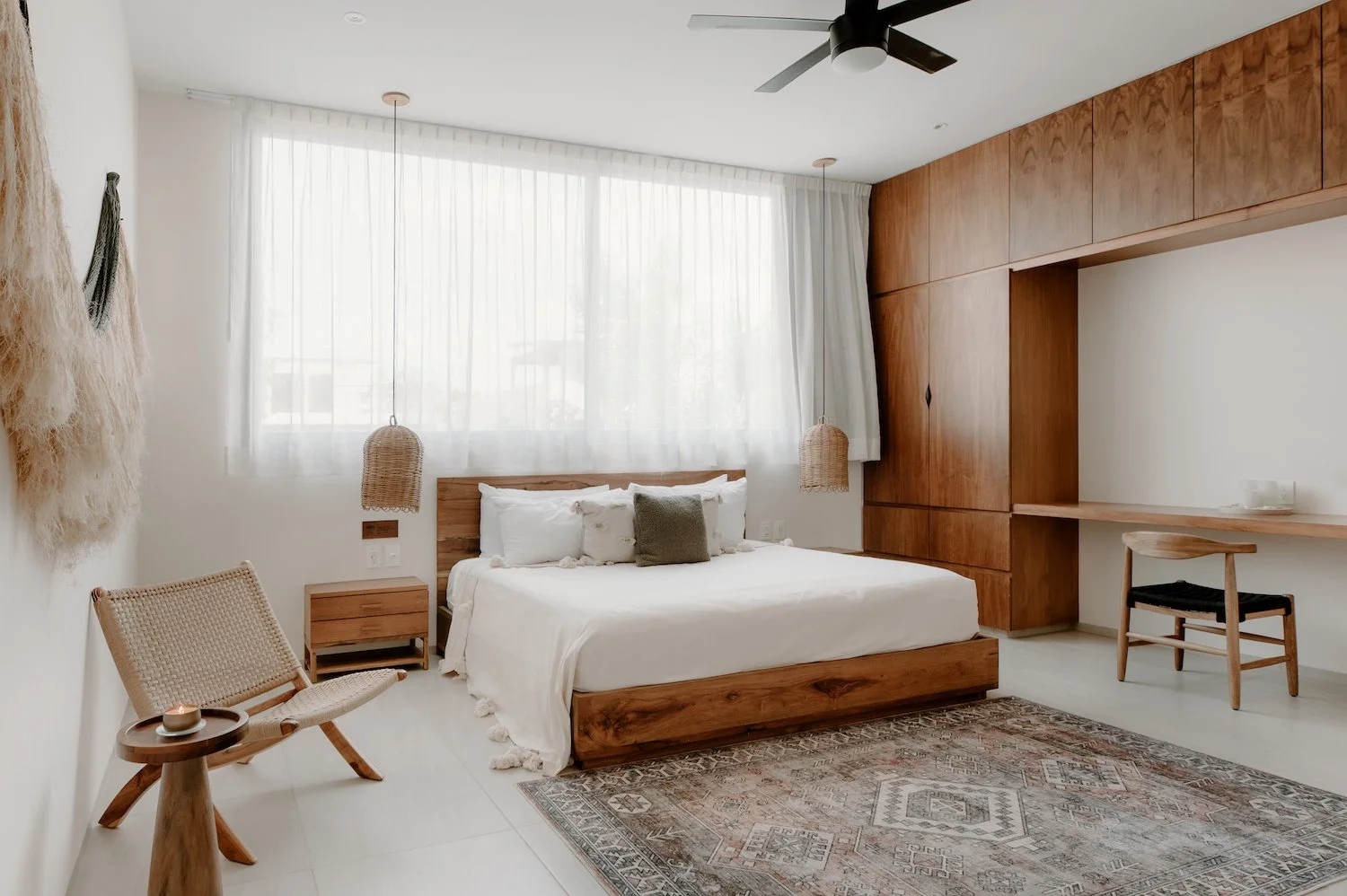A modern bedroom with a wooden bed frame, white bedding, and throw pillows. There are two hanging pendant lights, a small side table, a chair, and a wooden desk. The room has large windows with sheer curtains, a patterned rug, and wood-paneled walls.