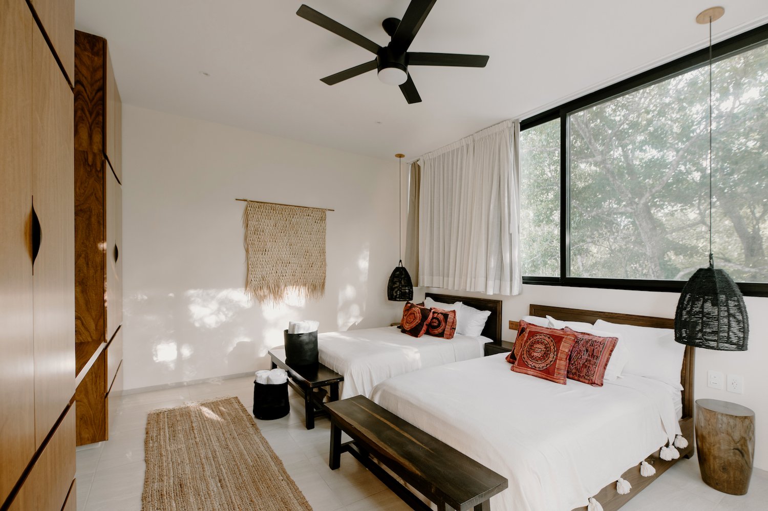 A bright bedroom with two beds, each with white linens and decorative pillows, a large window with white curtains, a wooden wardrobe on the left, woven wall decor, and modern pendant lighting, ceiling fan, and a rug on the floor.