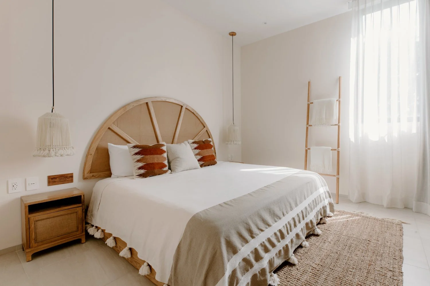 Minimalist bedroom with a large bed, woven headboard, white bedding, decorative pillows, hanging pendant lights, wooden side table, woven rug, and a wooden ladder with towels near a window with sheer curtains.