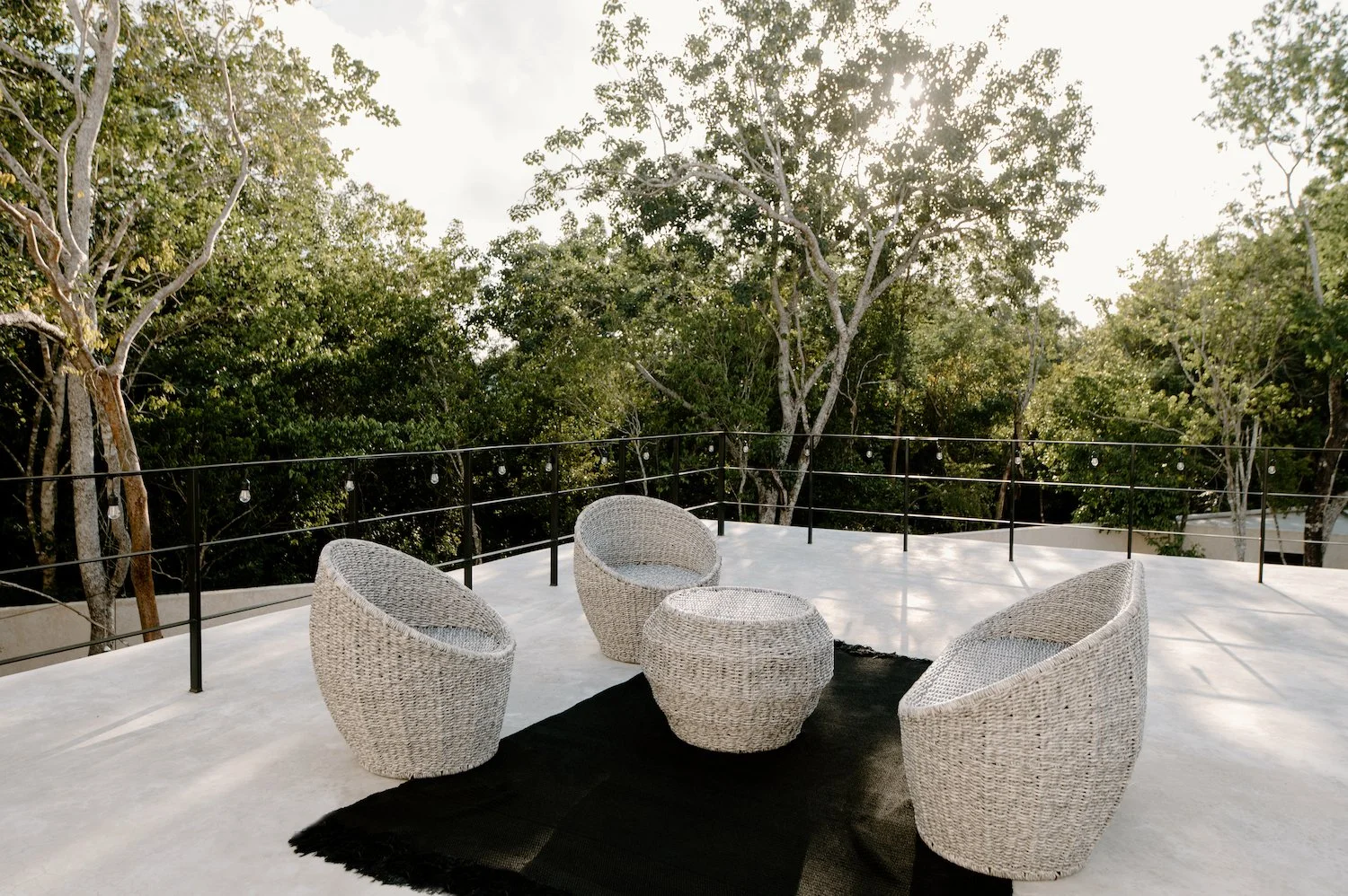 Outdoor patio with three wicker chairs and a small round wicker table on a black rug, surrounded by a black metal railing and lush green trees in the background.