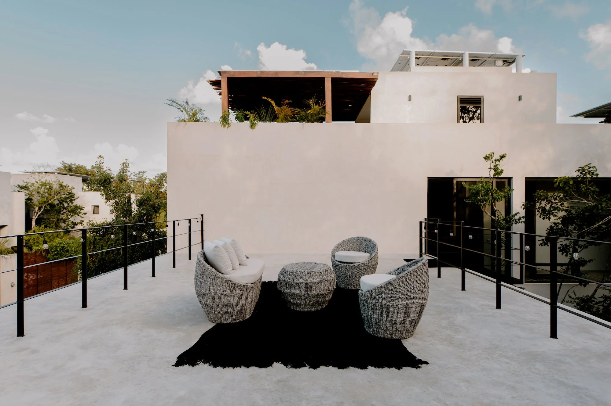 Outdoor balcony with four wicker chairs and a matching table on a black rug, surrounded by a black metal railing, overlooking a neighborhood with trees and modern white buildings under a partly cloudy sky.