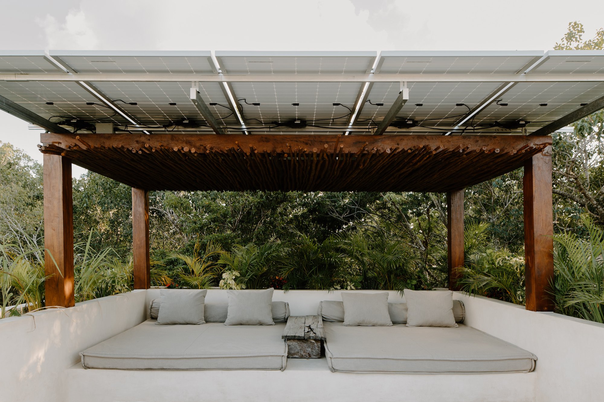 Outdoor seating area with a white sectional sofa with cushions, a rustic coffee table, and a wooden canopy with solar panels overhead, surrounded by lush greenery.