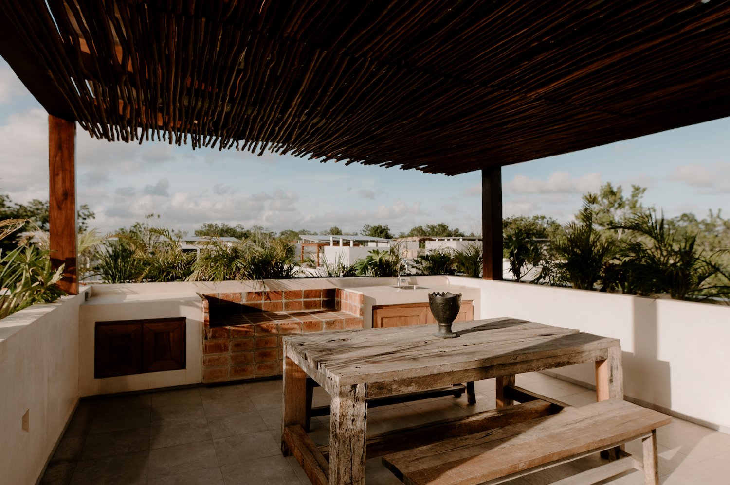 Outdoor terrace with a rustic wooden table and bench, brick and wood outdoor kitchen area with a sink, surrounded by green plants, under a wooden slat roof, with a view of the sky and trees.