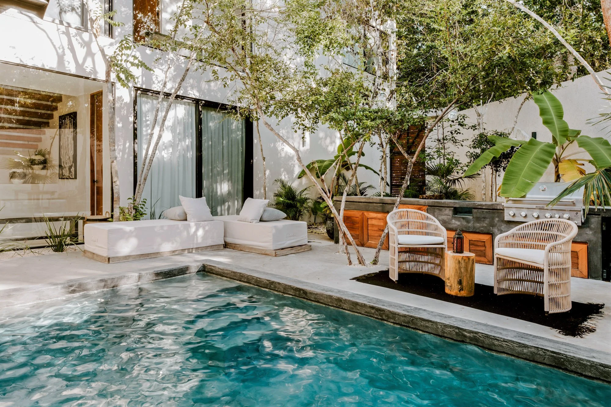 Outdoor patio area with a swimming pool, white lounge furniture, wooden chairs, green plants, and trees, with a white house in the background.