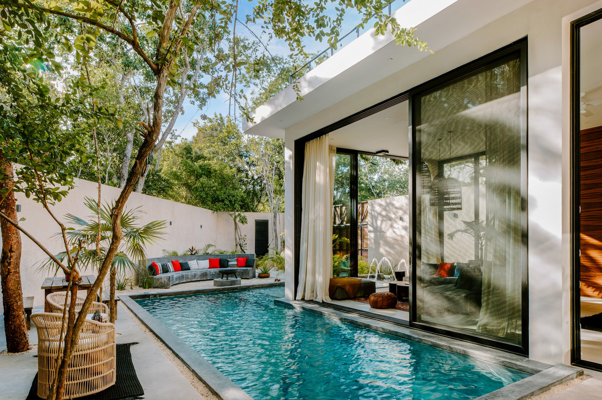 Modern house with glass sliding doors opening to a backyard with a swimming pool, outdoor seating, trees, and lush greenery.