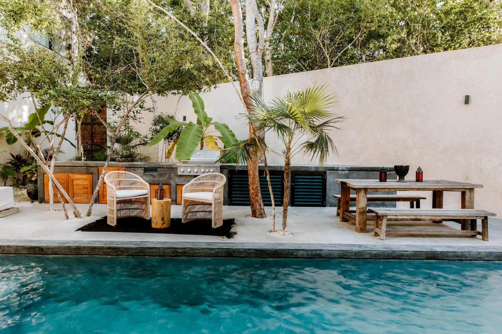 Outdoor poolside patio with wicker chairs, a wooden table, tropical plants, and a built-in grill area with a concrete countertop, enclosed by a tall beige wall and trees.