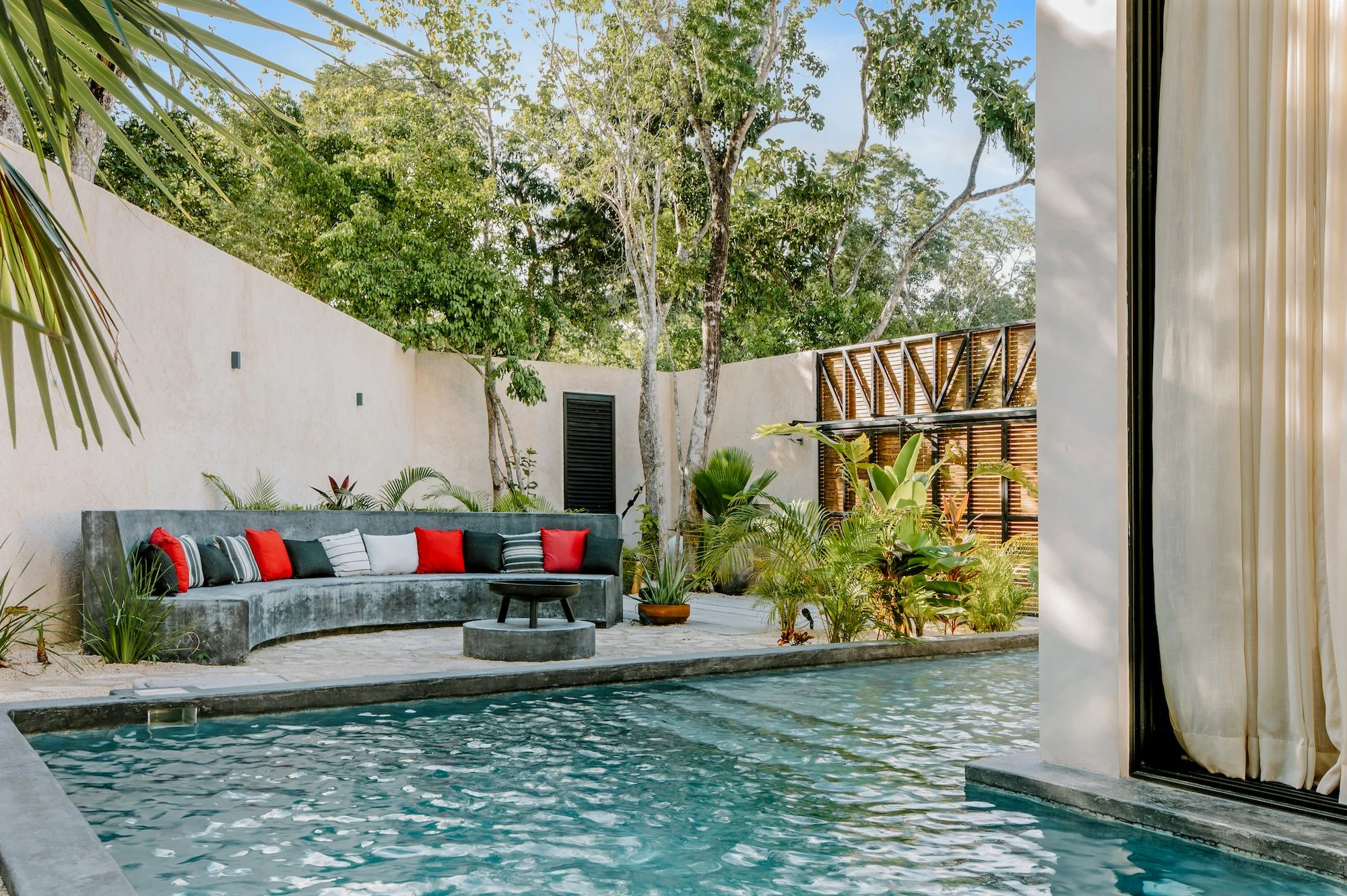 An outdoor pool with a curved concrete edge and clear water, surrounded by lush tropical plants. A curved gray concrete bench with red and black striped pillows sits against a beige wall. Tall trees and a wooden fence are visible in the background, with the sky partly cloudy.