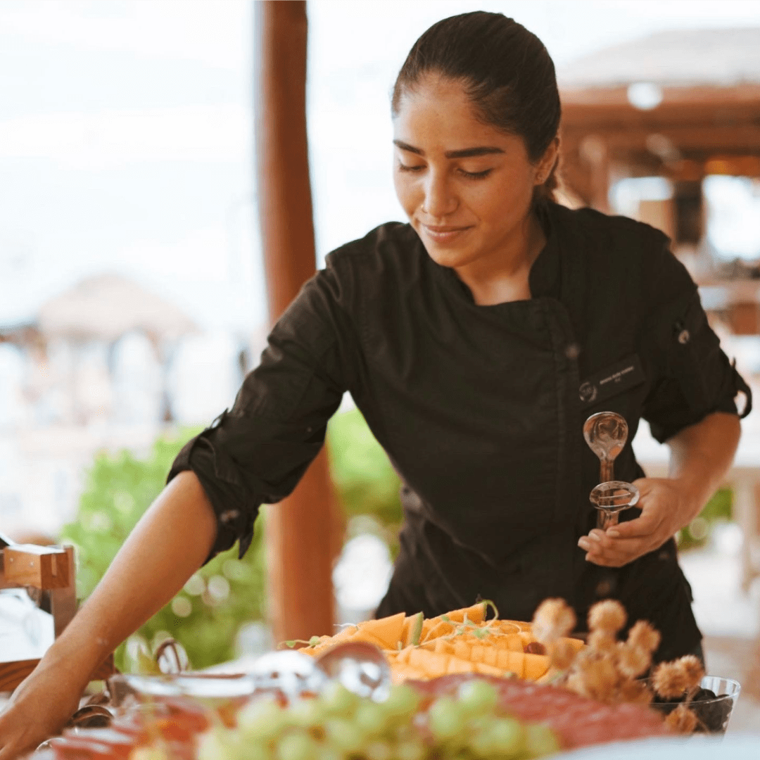 A woman in a black chef's coat preparing a fruit platter with melons, grapes, and other fresh fruits at an outdoor setting.