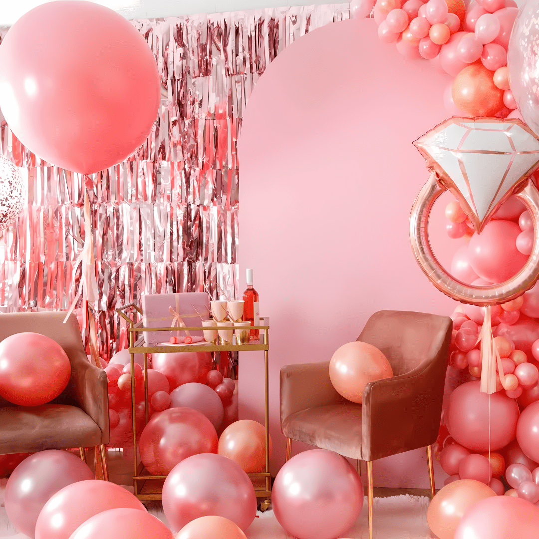 A pink-themed party setup with balloons, a balloon arch, a pink background wall, a champagne glow candle, a pink drink, and chairs.