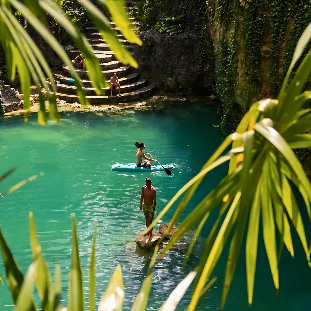 People swimming and sitting on rocks around a turquoise natural pool, seen through green palm leaves.