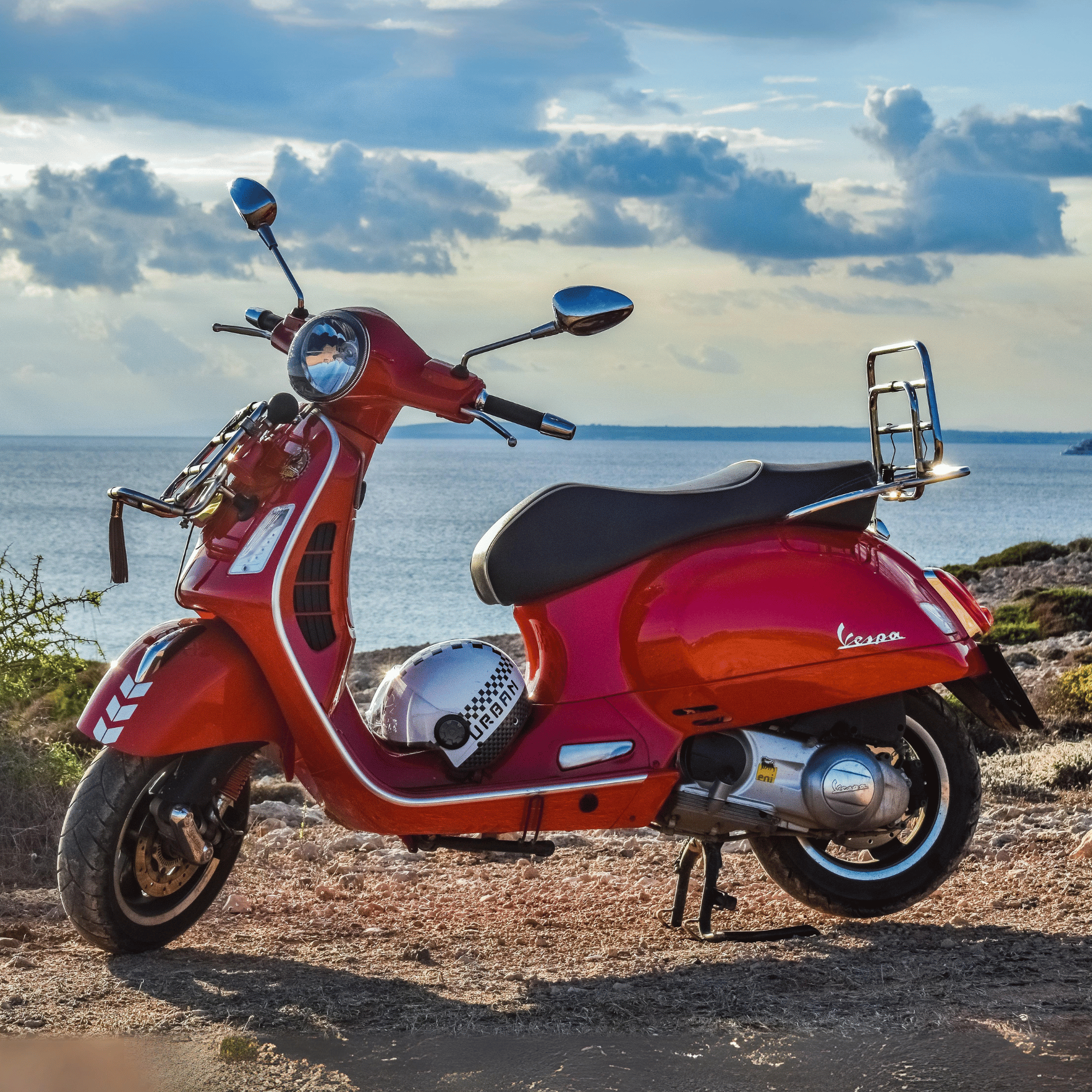 Red Vespa scooter with side mirrors and GPS mounted on the handlebar, parked on a rocky surface near a body of water with cloudy sky in the background.