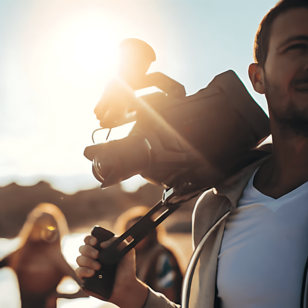 A man smiling outdoors during sunset, carrying a child on his shoulder, with sunlight creating a backlit effect, and other people in the background near a body of water.