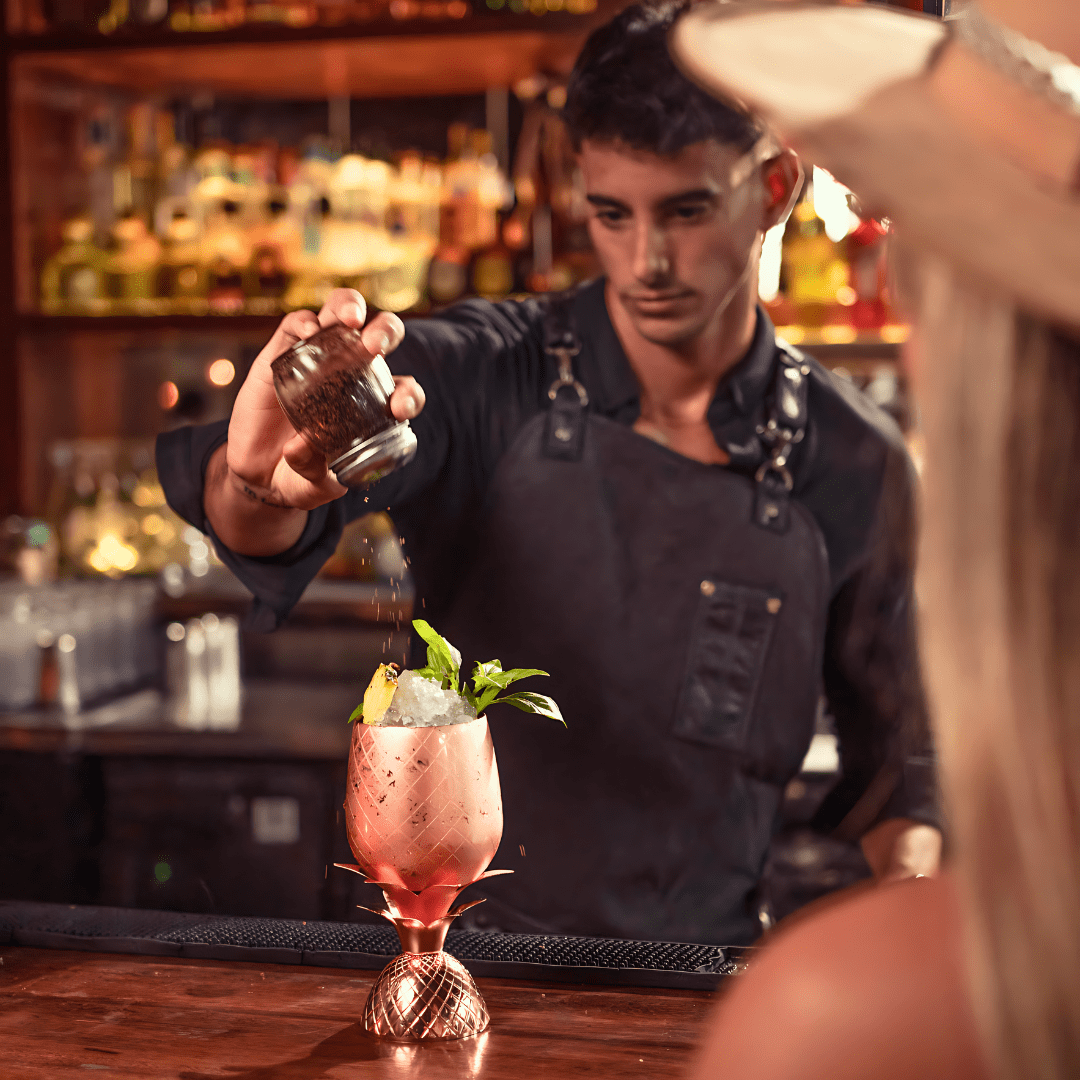 Bartender pouring spice or seasoning onto a cocktail in a copper pineapple-shaped glass at a bar.