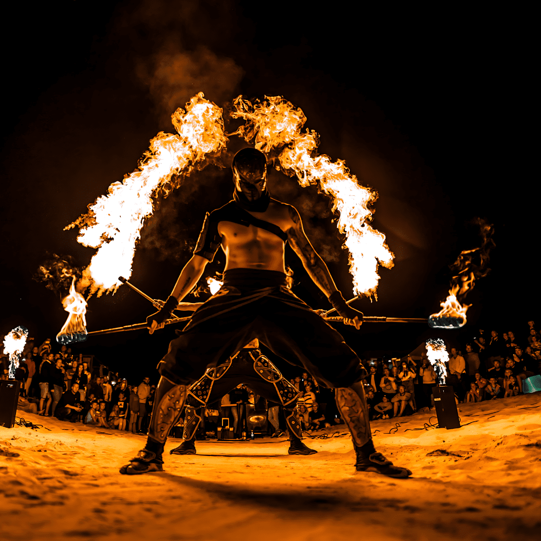 A performer in dark clothing and face paint controls fire on two staffs during a nighttime fire show, with a large audience watching.