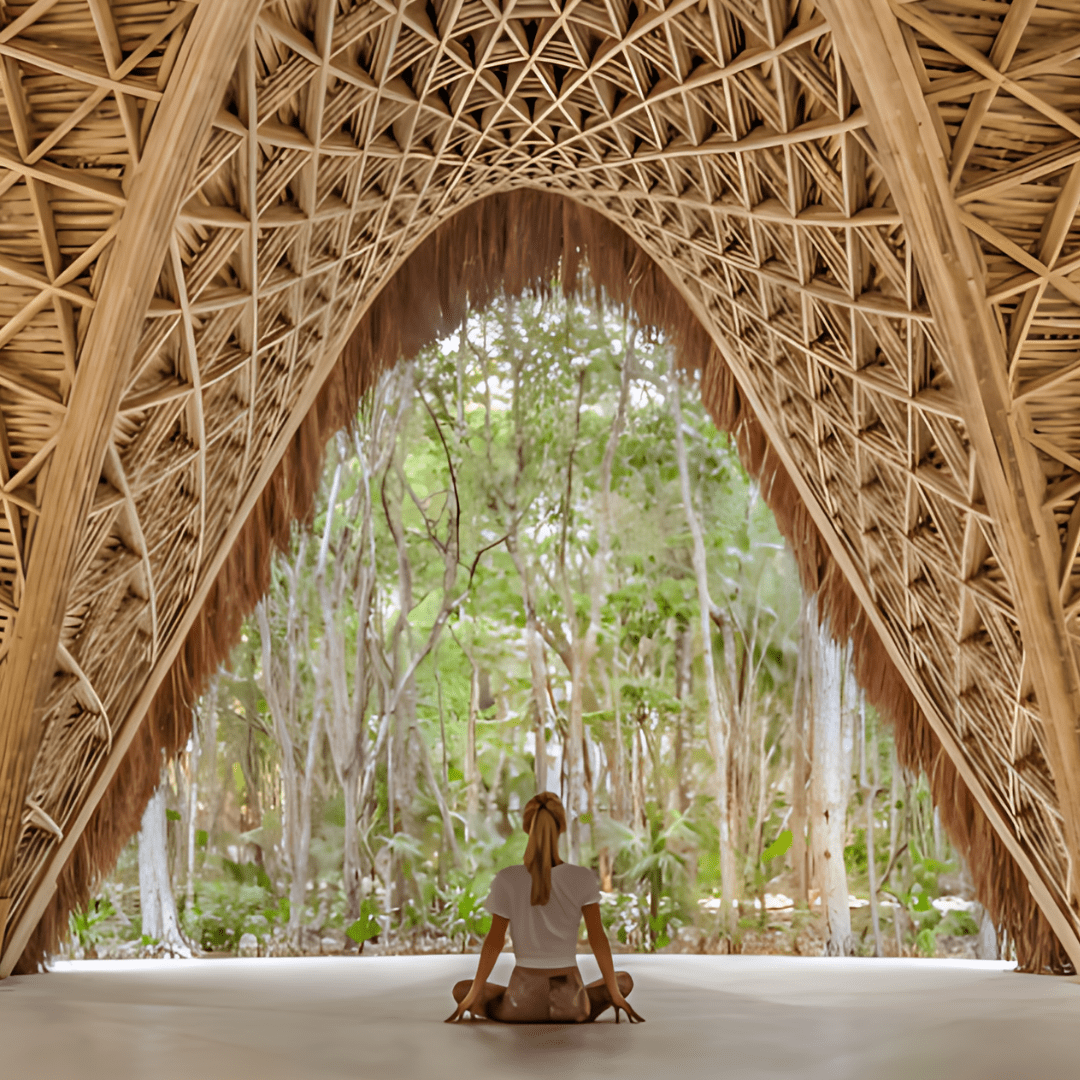 A person practicing yoga or meditation in a natural, outdoor setting surrounded by trees, viewed from inside a woven, thatched-roof hut or tent.