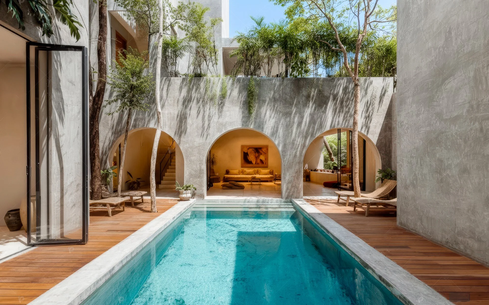 Indoor swimming pool with wooden deck, surrounded by modern concrete walls with trees growing through openings, and a cozy indoor lounge area visible through an archway.