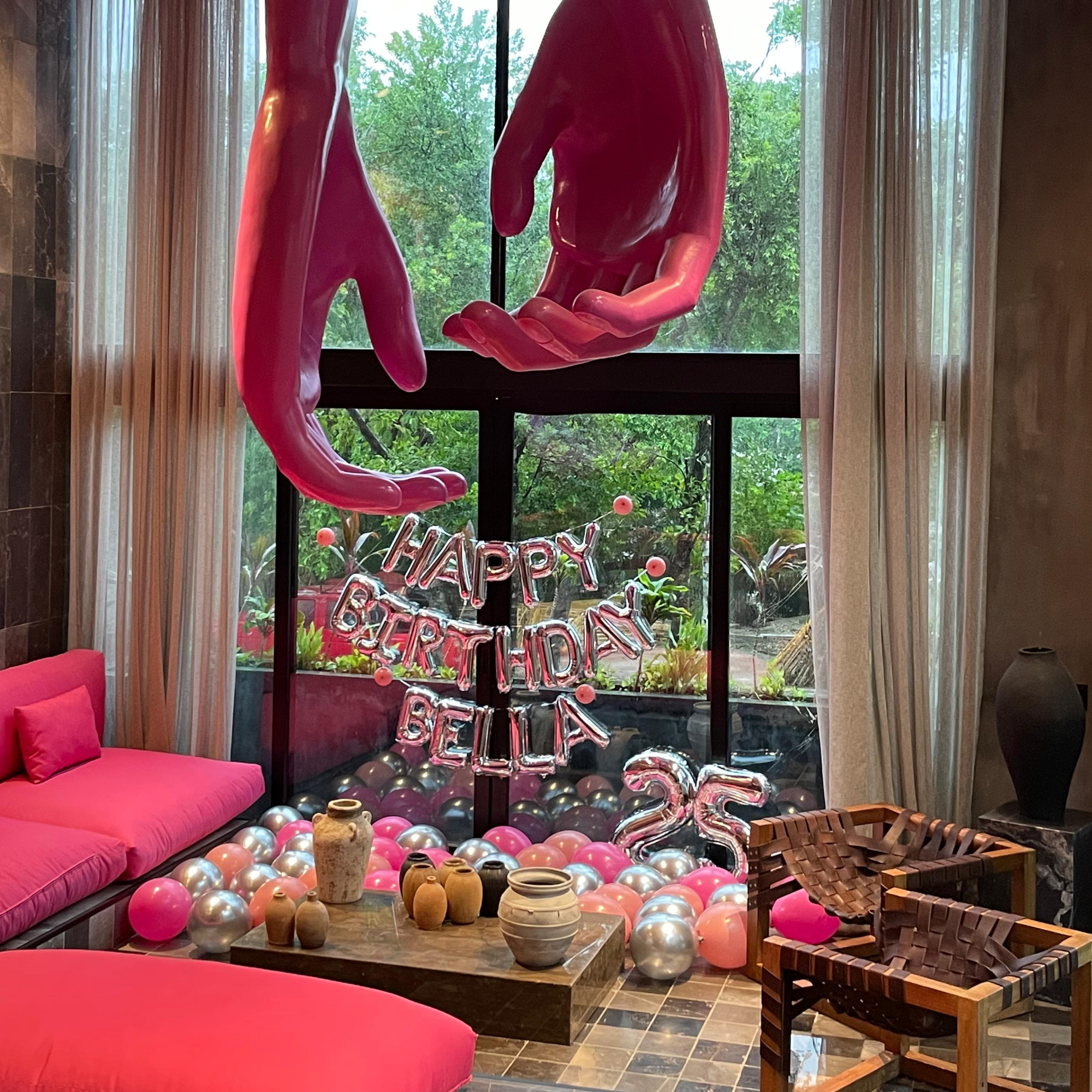 Interior of a room decorated for a celebration, featuring large pink sculptures of hands, a neon "Happy Birthday" sign, balloons scattered on the floor, a pink cushioned bench, and various ceramic pots and vases on tables.