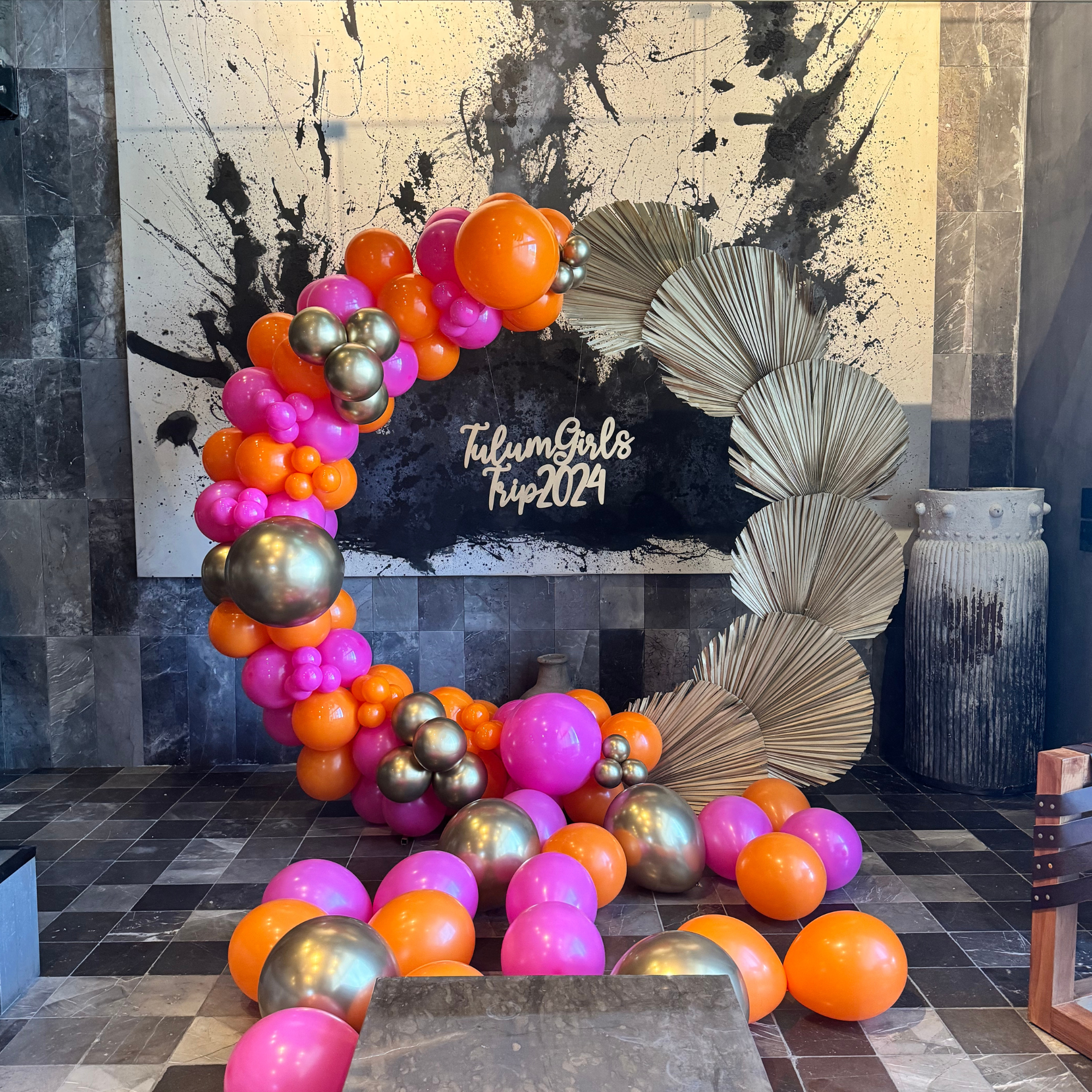 Colorful balloon arrangement with pink, orange, silver, and purple balloons, set against a white and black splash art backdrop with the words "Tulumi Girls Trip 2024" at the bottom, on a tiled floor.